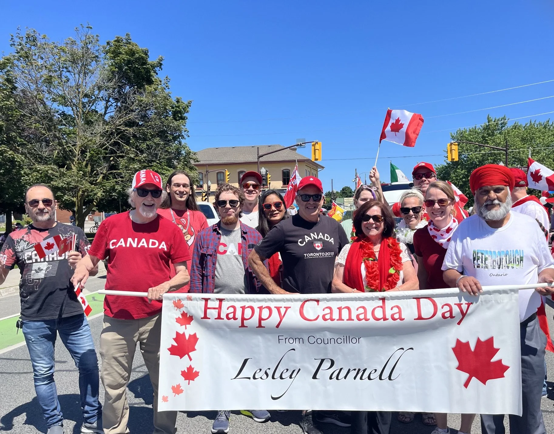 Group of people participating in a Canada Day parade, holding a banner that says "Happy Canada Day" with red maple leaf symbols. They are wearing red clothing and accessories, some waving Canadian flags, on a sunny day with clear blue skies.