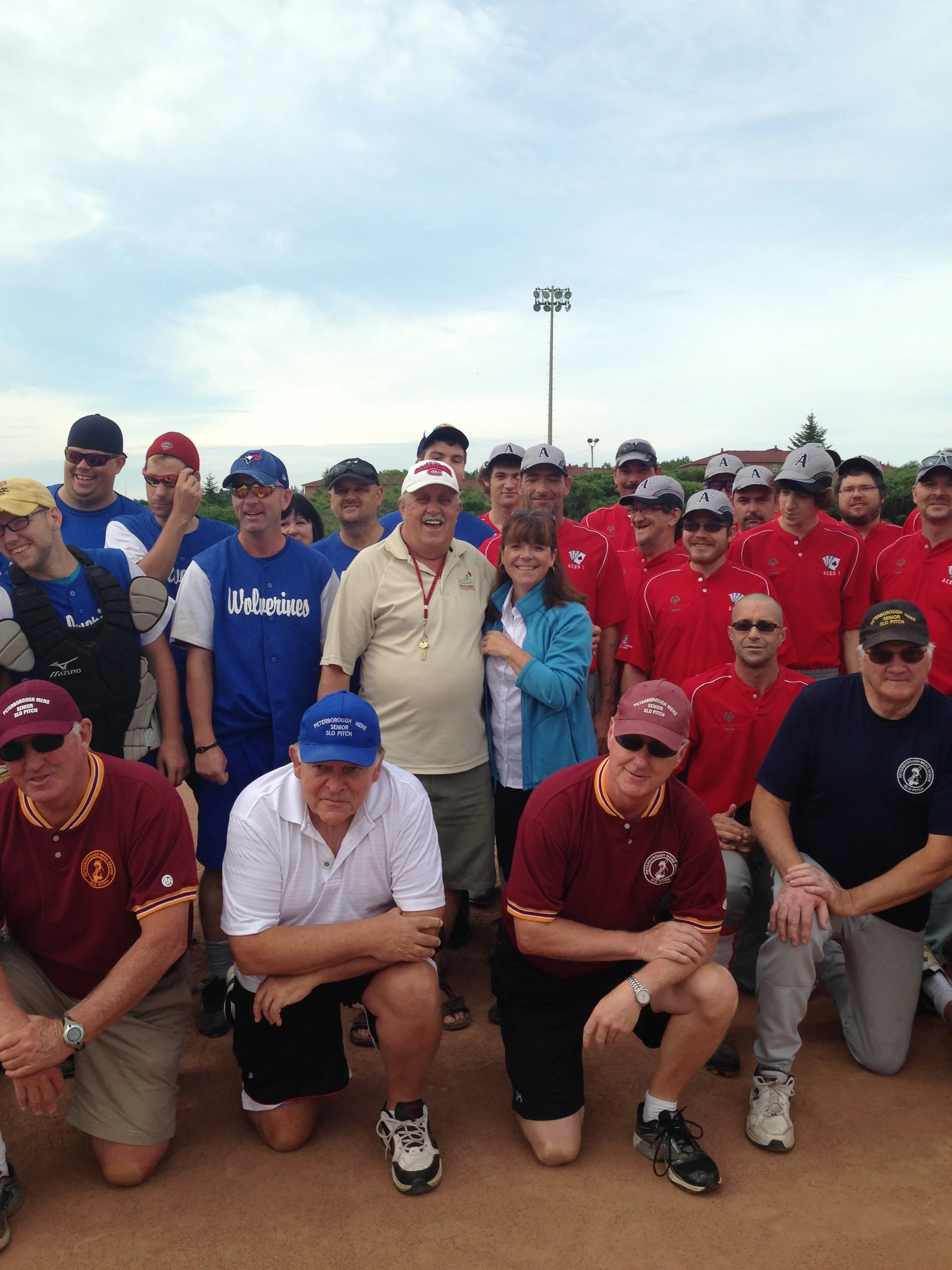 A group of people outdoors, some kneeling and some standing, posing for a photo. They are wearing sports or team shirts, hats, and sunglasses. The background features a cloudy sky, a tall light pole, and some trees and buildings.