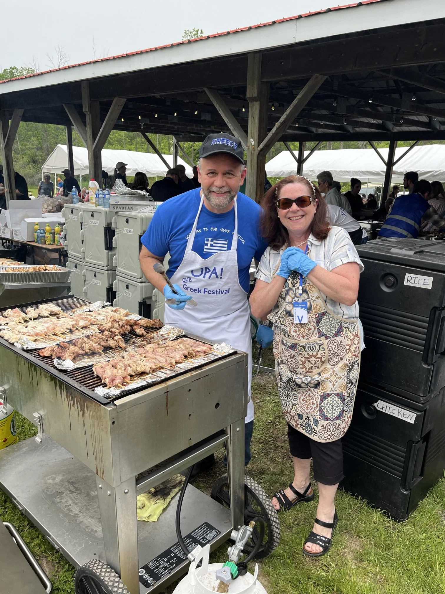 Two smiling people, a man in a blue shirt and apron and a woman in sunglasses, standing at a grill with skewered meat, under a pavilion at an outdoor Greek festival. Background shows a crowd and picnic tents.