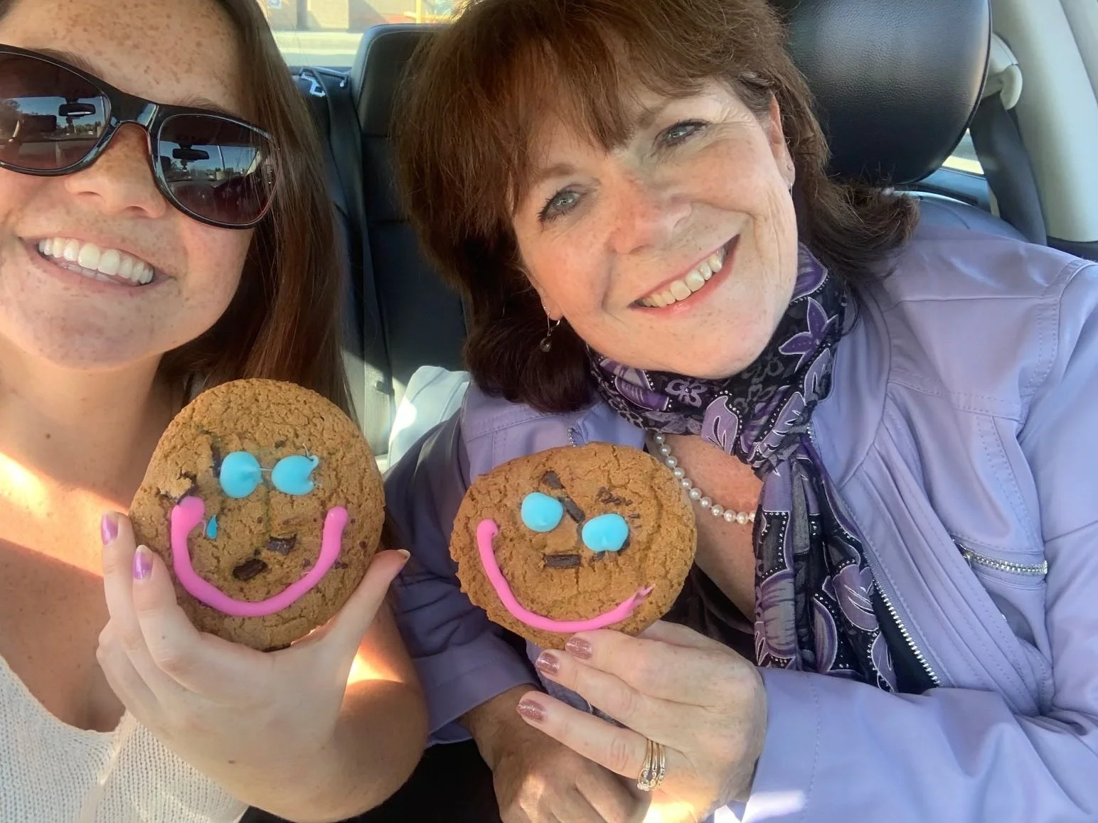 Two women sitting in a car holding decorated cookies. One woman is wearing sunglasses and a white top, the other wearing a purple jacket and a scarf. Both cookies are decorated with colorful icing in the shape of smiling faces.