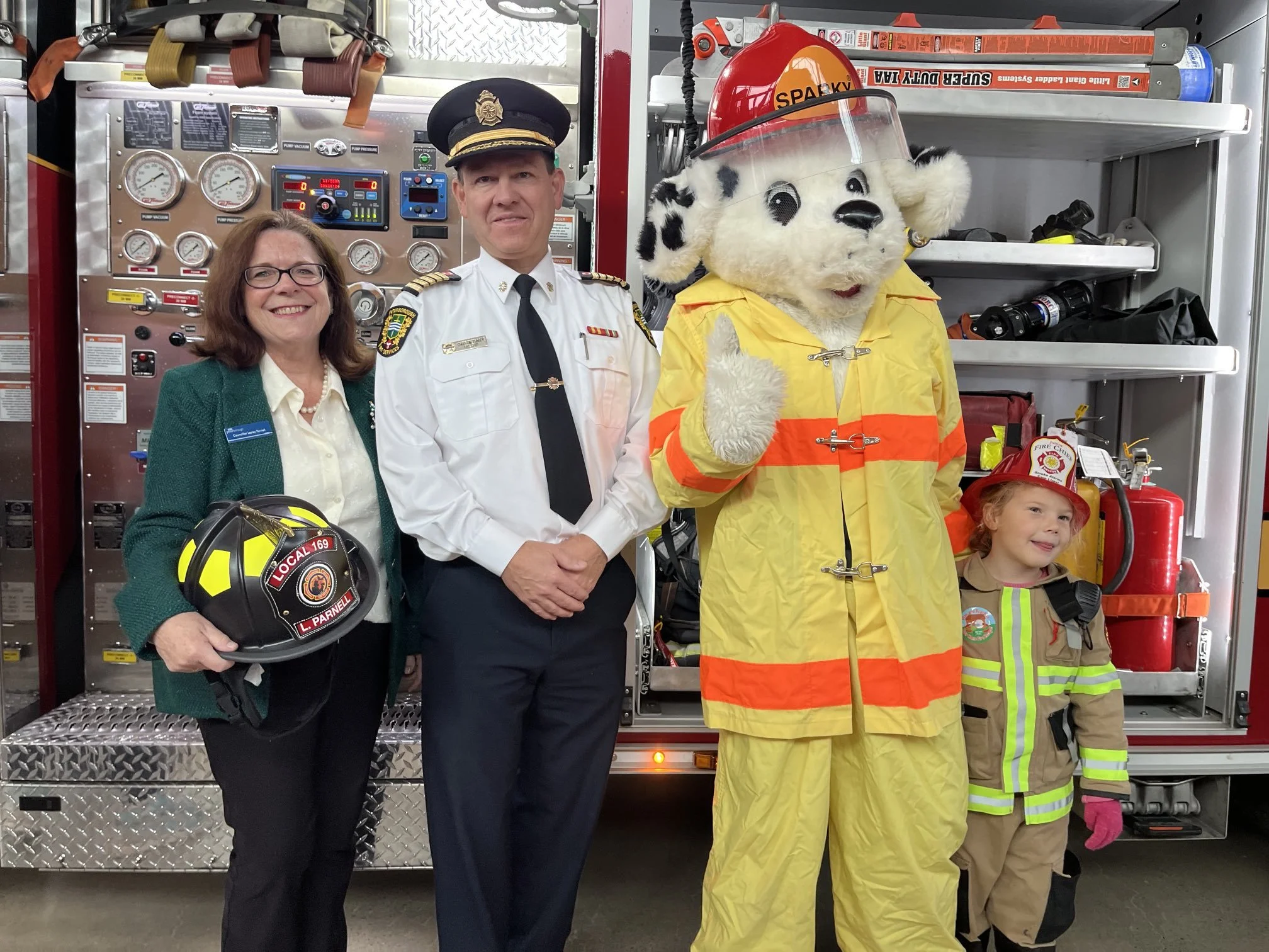 A firefighter holding a helmet, a woman with glasses and a badge, a firefighter in uniform, a mascot dressed as a dog in firefighting gear, and a young girl in firefighter uniform standing in front of a fire truck.