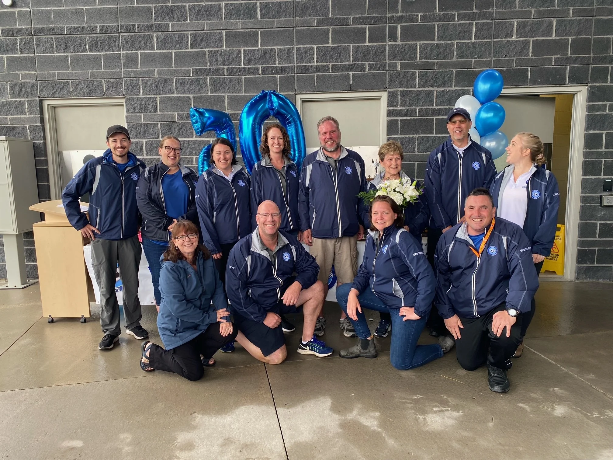 A group of people, including men and women, standing and kneeling in front of gray brick wall decoration with blue balloons and large blue foil balloons forming the number 20. They are dressed in matching navy jackets with a logo, celebrating an even
