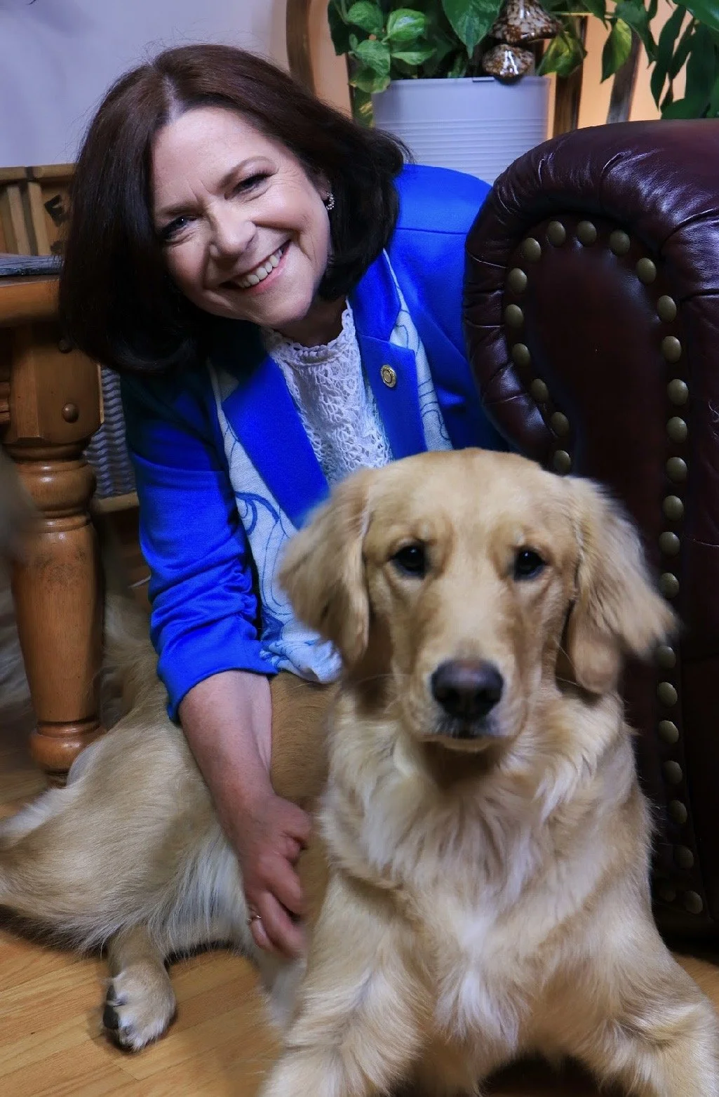 A woman with shoulder-length dark hair and a bright smile, wearing a blue blazer and lace top, is sitting on the floor with a golden retriever dog, surrounded by wooden and leather furniture and green plants in the background.