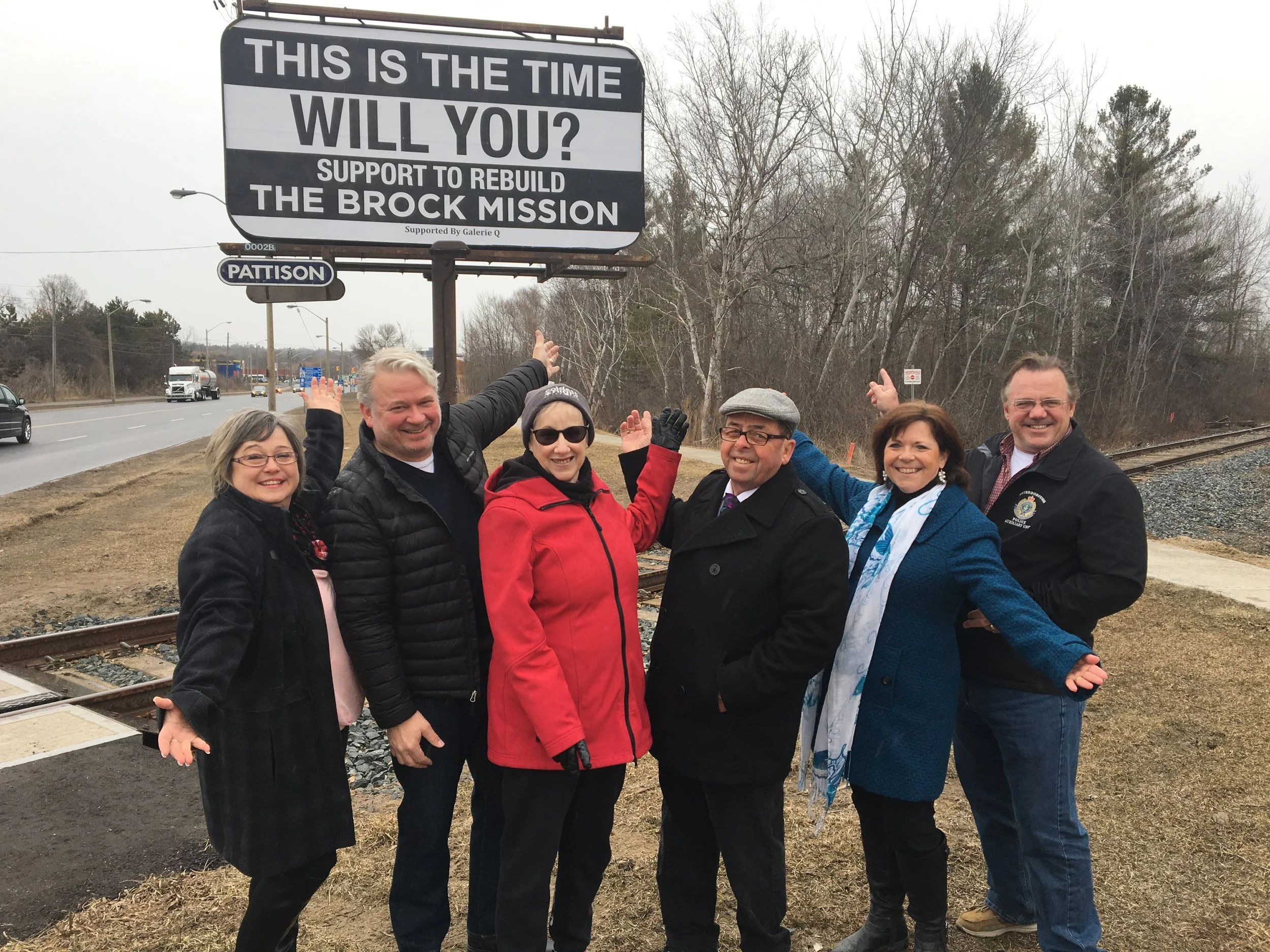 A group of six smiling people standing near railroad tracks holding a sign that reads, 'This is the time will you? Support to rebuild the Brock mission.' They are outdoors, dressed in jackets, with trees and a road in the background.