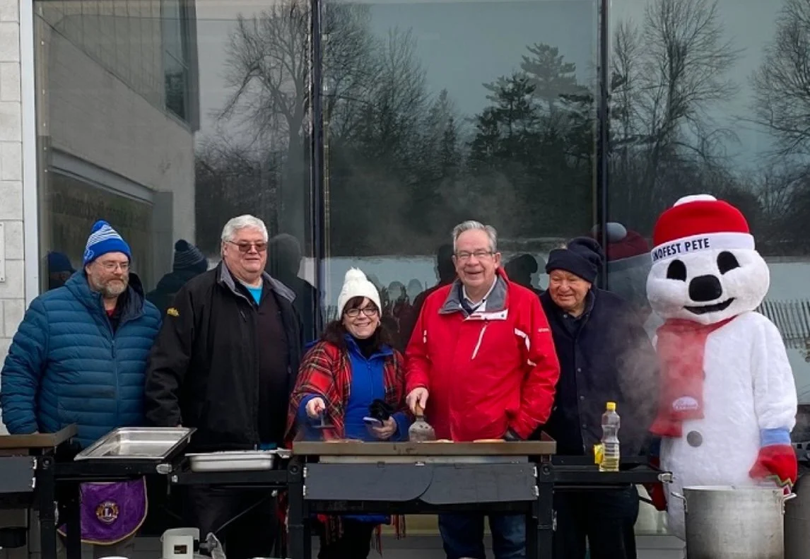 Five people and a snowman mascot standing outside in front of a building with large glass windows. The people are dressed in winter clothing and appear to be participating in an outdoor event during cold weather.