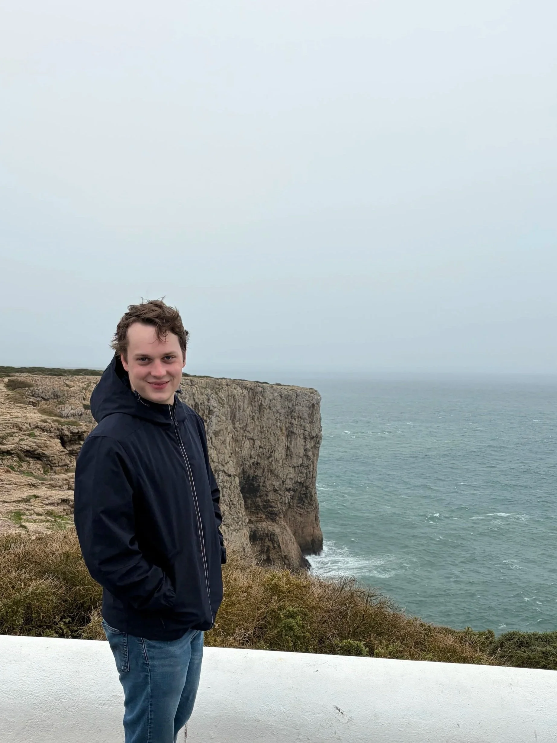 A young man with curly brown hair wearing a black jacket and blue jeans standing near white railing on a coastal cliff with ocean and cloudy sky in the background.