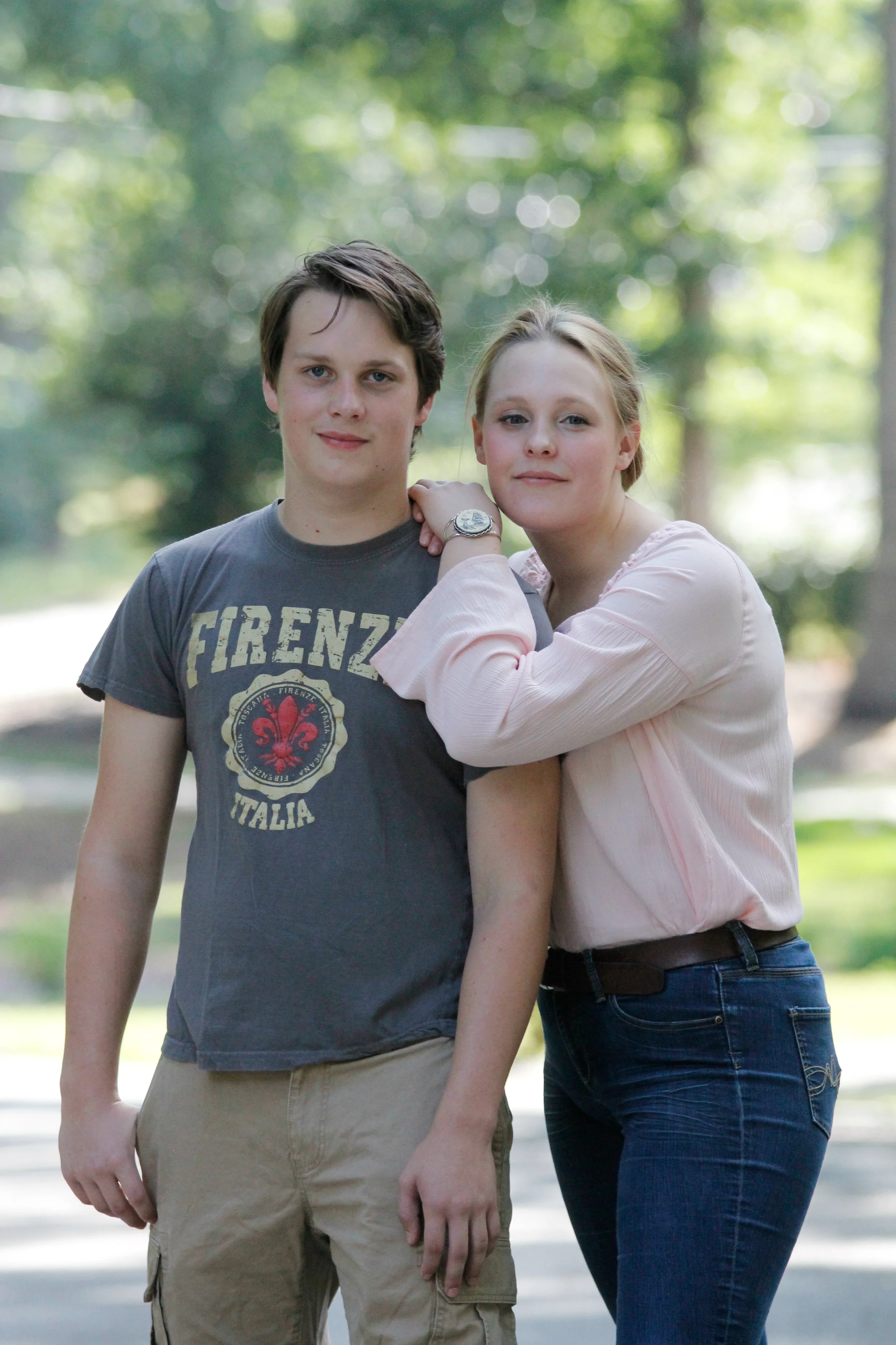 A young man and young woman standing outdoors with a blurred leafy background. The woman has her arm resting on the man's shoulder.