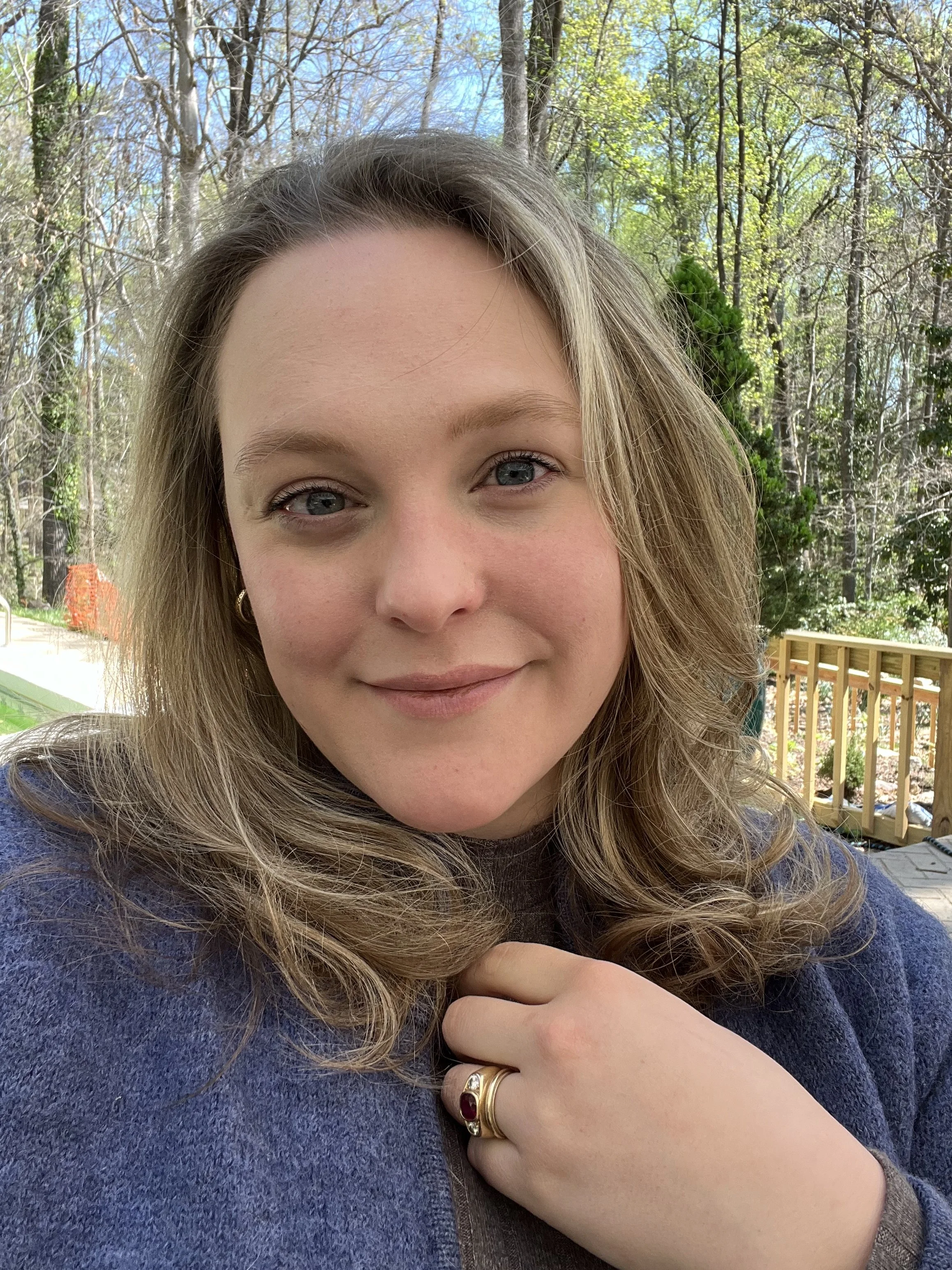 Close-up of a woman with blonde hair, blue eyes, and a subtle smile, wearing a gold ring and a dark sweater, with a background of trees and a wooden deck.