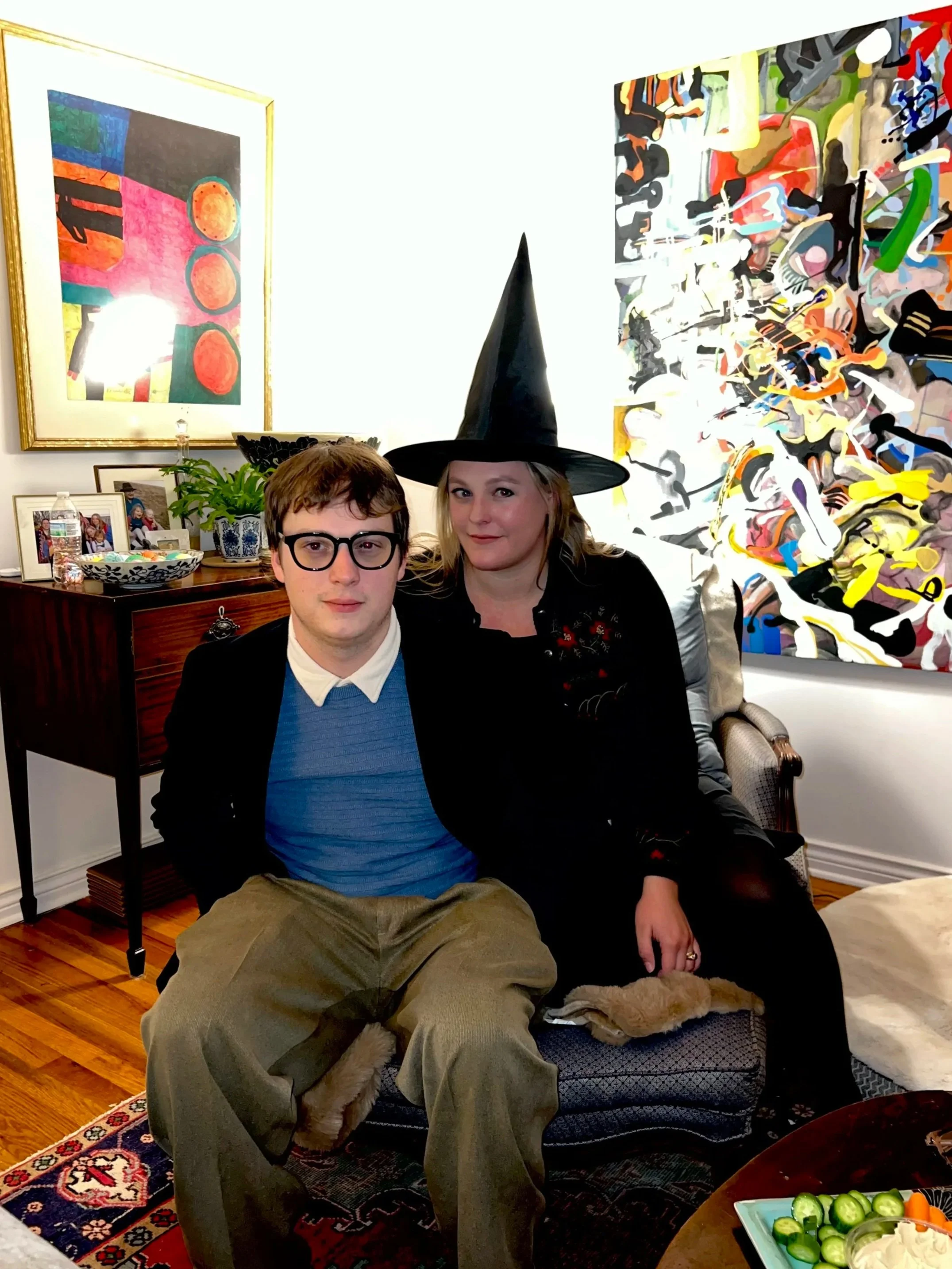 Two people sitting on a cushioned armchair in an art-filled living room, one wearing a witch hat. There are paintings on the wall and a wooden sideboard with pictures, a potted plant, and drinks. A table with sliced cucumbers and vegetables is visible in the foreground.