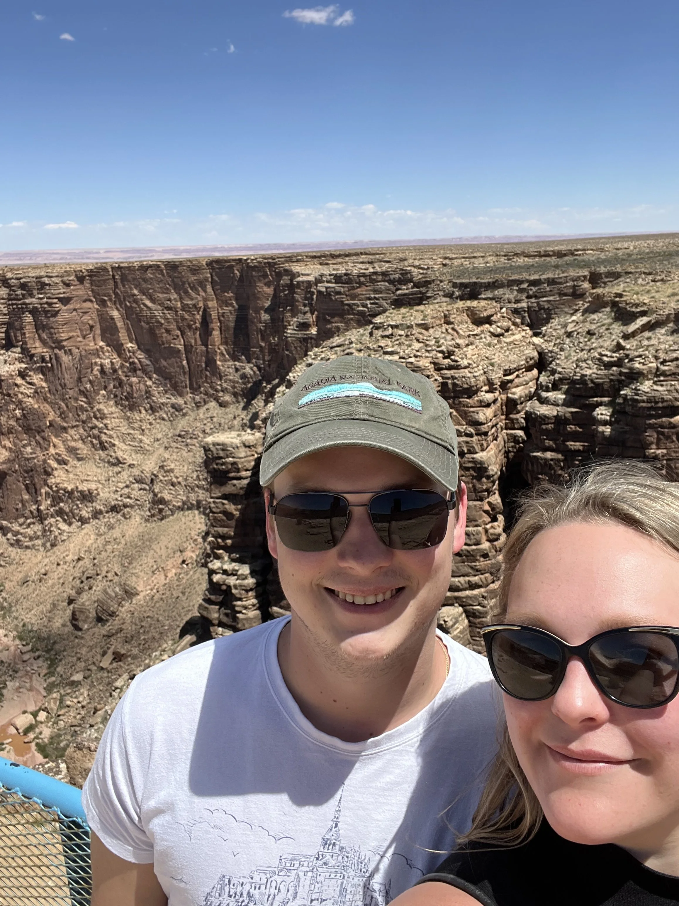 Two people taking a selfie at the Grand Canyon, with the canyon's rocky cliffs and a blue sky in the background.