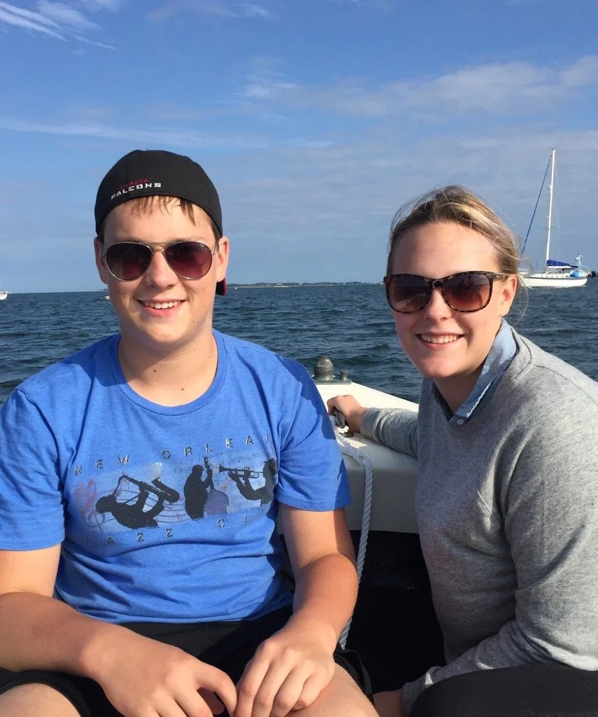 Two young people smiling and sitting on a boat with water and sailboats in the background on a sunny day.