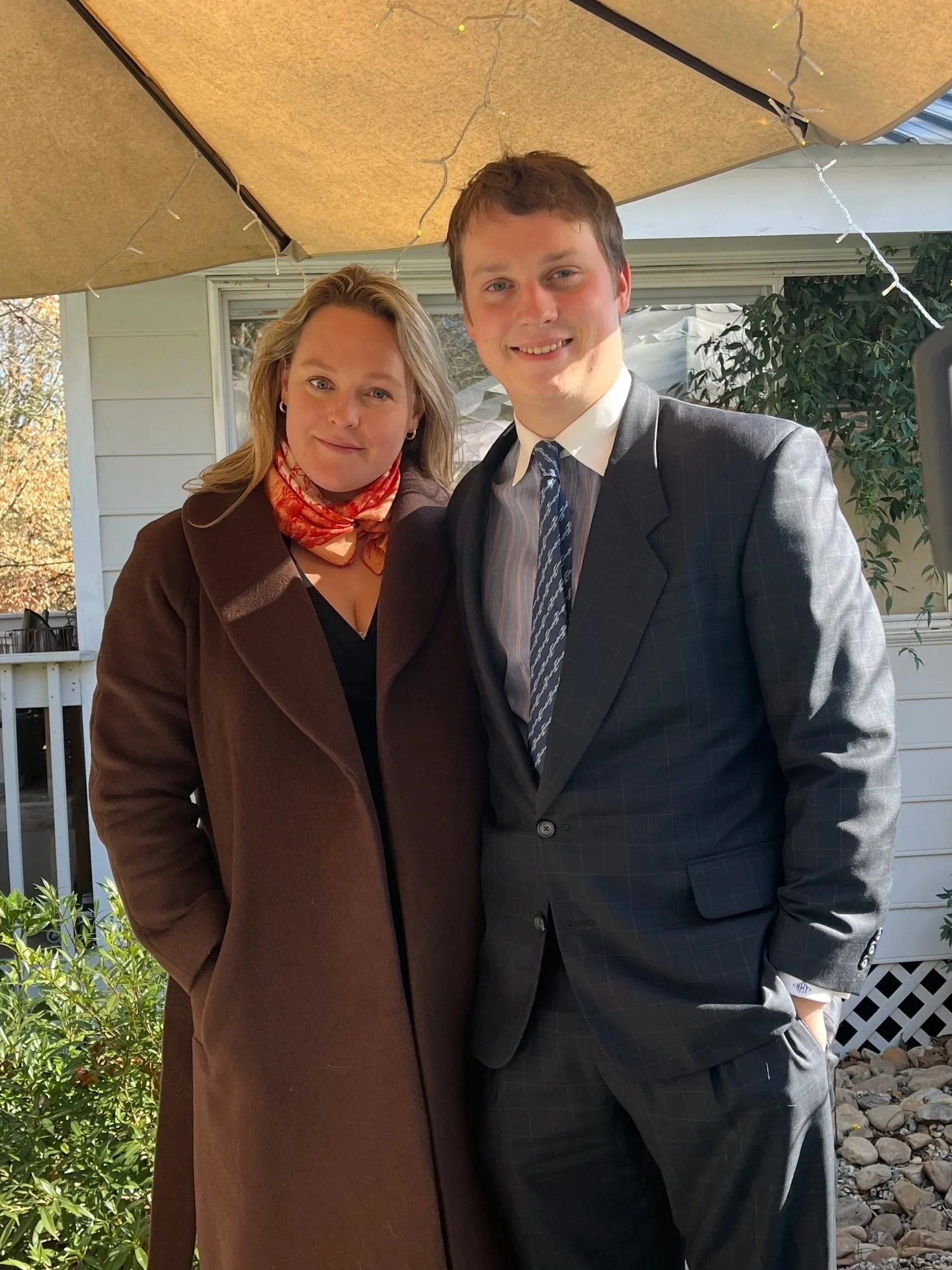 A man and woman dressed in formal attire standing side by side outside near a house, with a brown umbrella overhead. The woman wears a brown coat and a patterned scarf, and the man wears a gray checkered suit with a striped tie. They are smiling and looking at the camera.