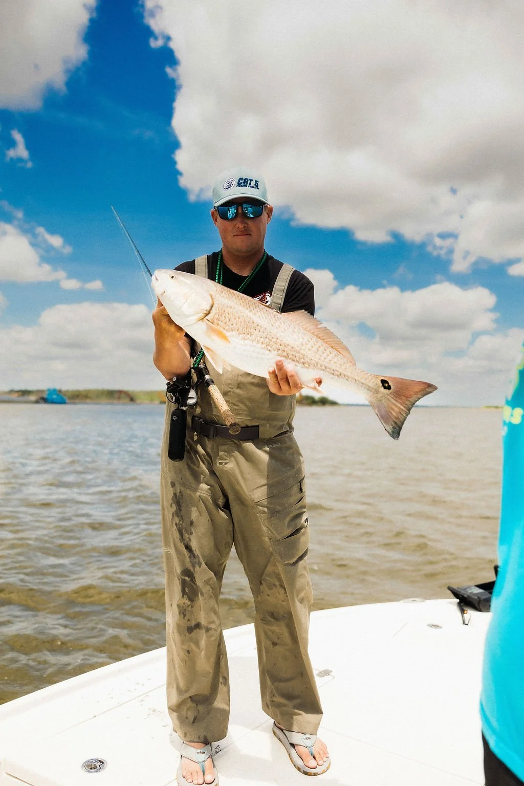 Man in sunglasses and fishing gear holding a large fish on a boat, with water and cloudy sky in the background.