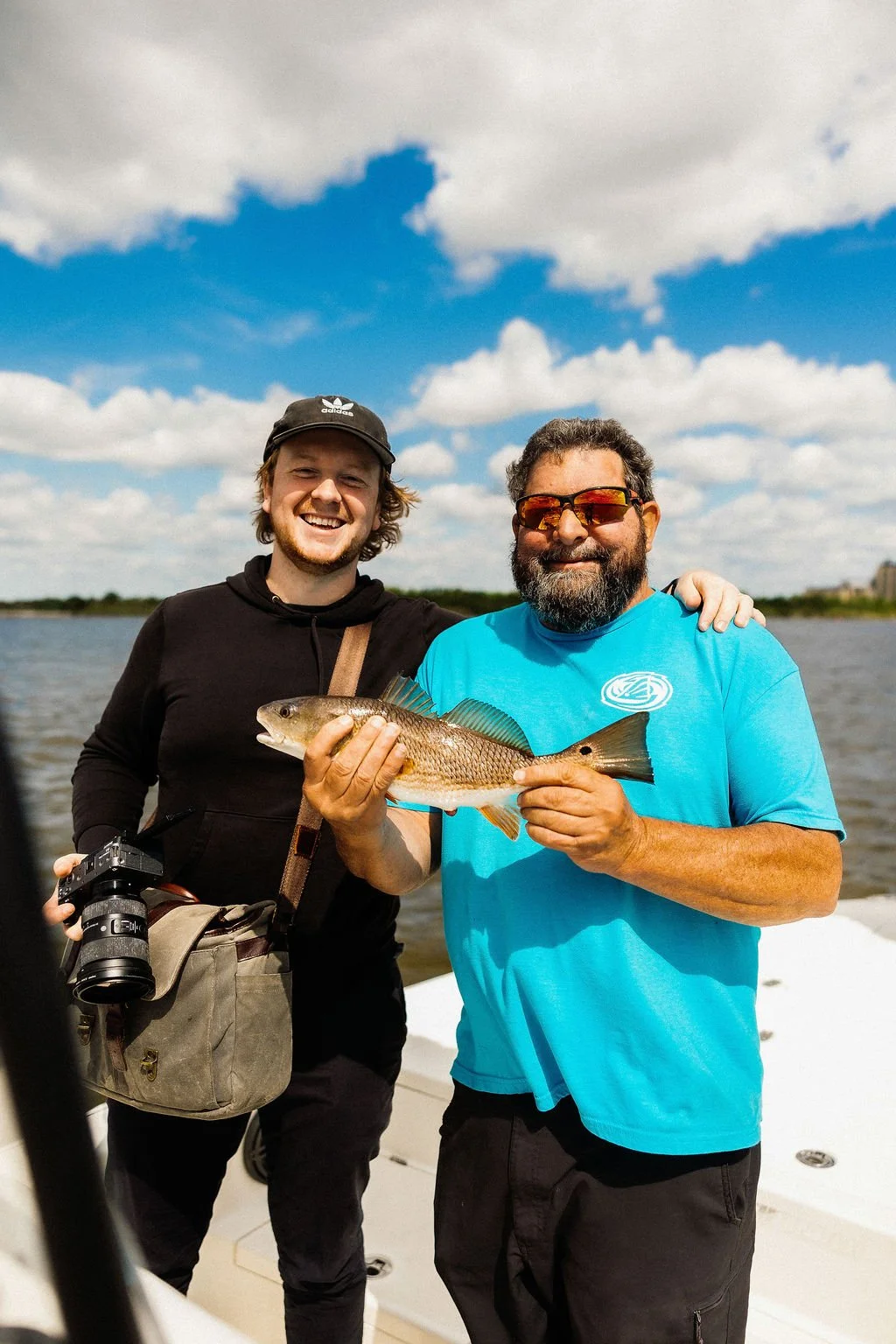 Two men smiling on a boat, one holding a fish, under a partly cloudy sky.