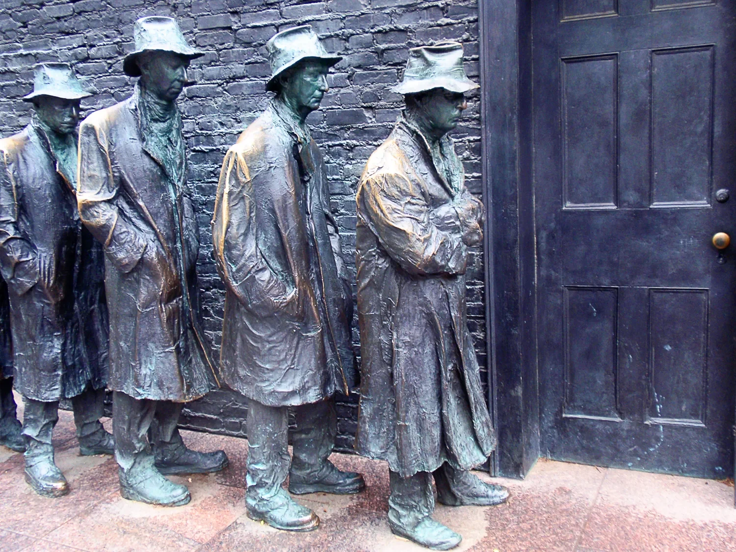 Sculpture of five gaunt figures wearing overcoats and hats standing single-file against a rough wall waiting for public food assistance.