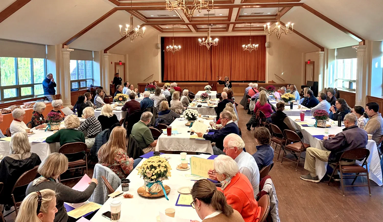 Attendees seated at tables listen to a speaker at a podium in a chandelier‑lit hall during a community gathering for Faith in Action at St. Thomas’ Church Whitemarsh.