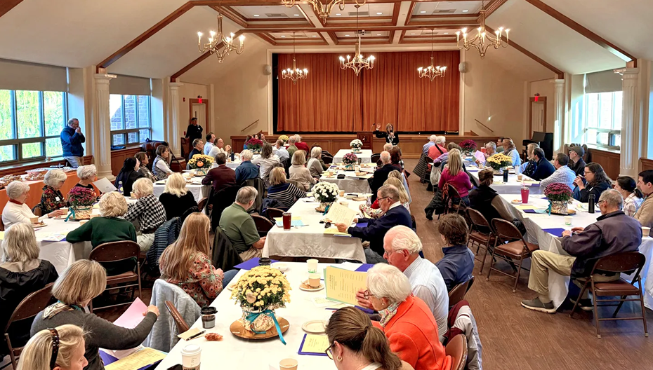Attendees seated at tables listen to a speaker at a podium in a chandelier‑lit hall during a community gathering for Faith in Action at St. Thomas’ Church Whitemarsh.