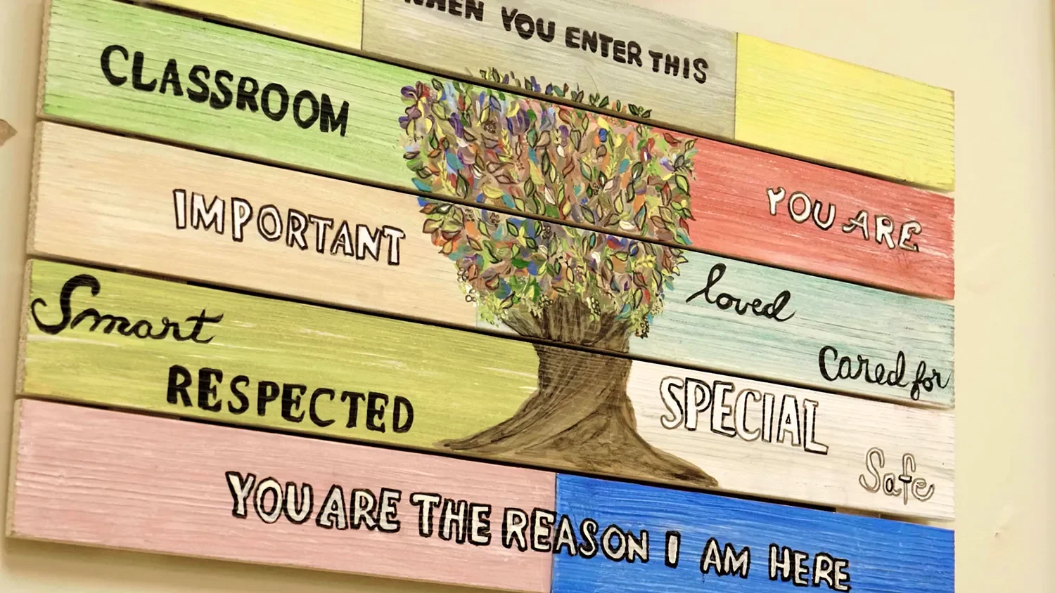 A colorful wooden classroom sign displays positive messages such as “You are important,” “Smart,” “Loved,” and “Safe,” centered around an illustrated tree at St. Thomas’  Preschool.