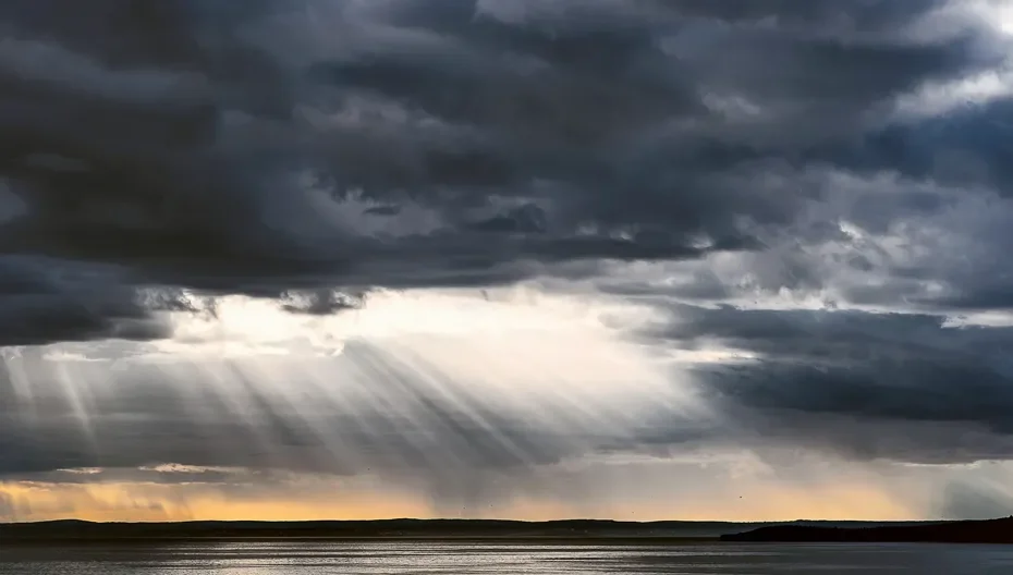 Sunlight breaks through dark and stormy clouds over a lake with mountains in the background.