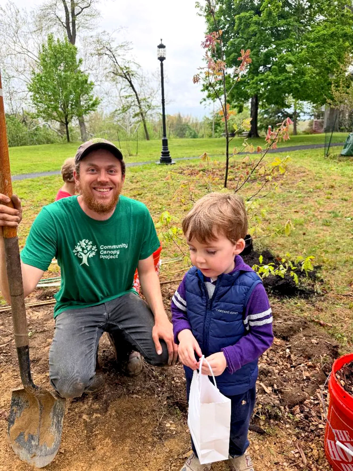 A man wearing a “Community Canopy Project” shirt helps a young child plant a small tree outside at St. Thomas's Preschool.