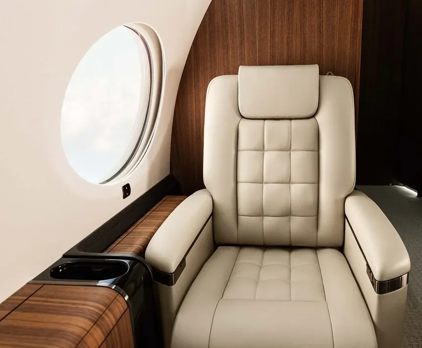 Inside an airplane cabin, there is a beige leather seat with a high backrest and adjustable headrest. Adjacent to the seat is a wooden panel with a built-in cup holder. A round airplane window is visible on the left side of the image.
