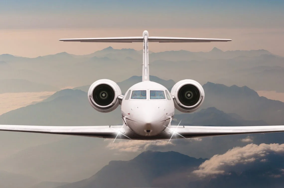 A front view of a white private jet flying above the mountains and clouds.