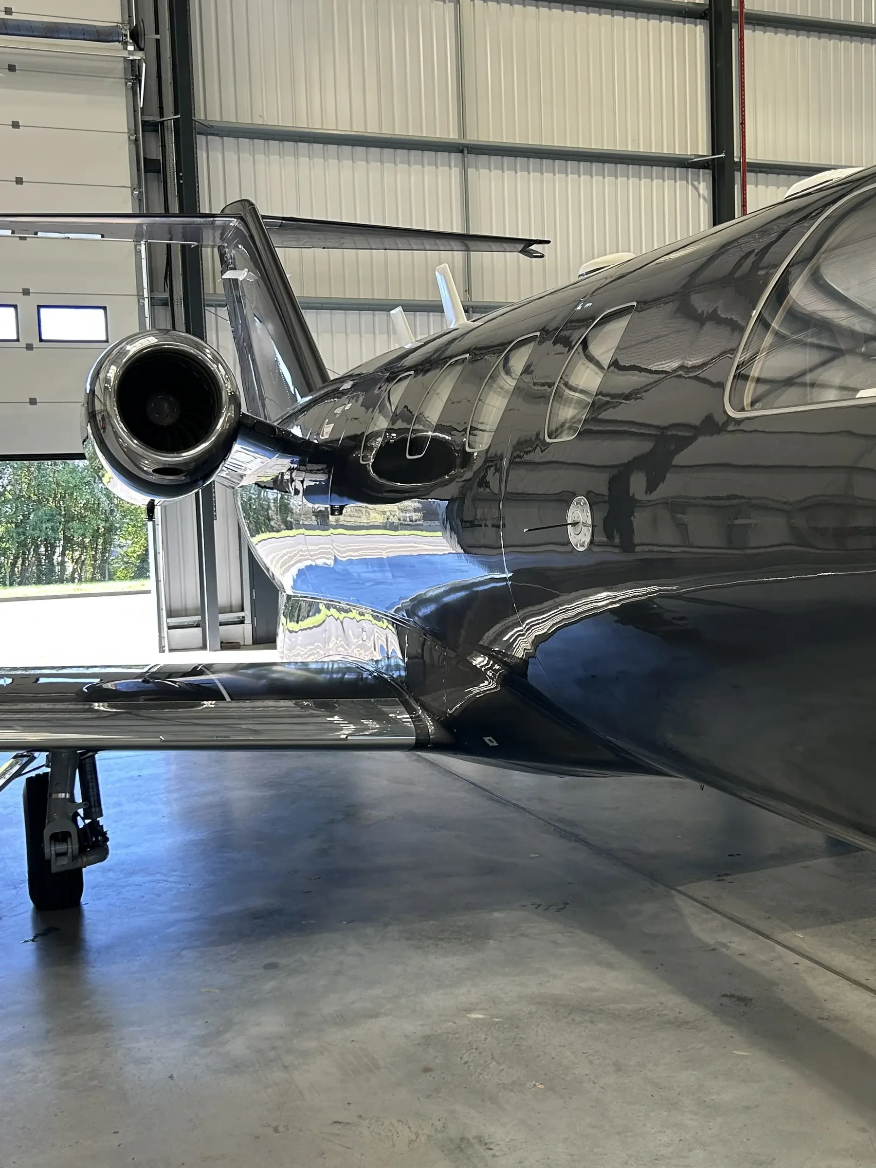 A black private jet with shiny exterior and a large jet engine on the side, parked inside a spacious garage with metal walls and a concrete floor.