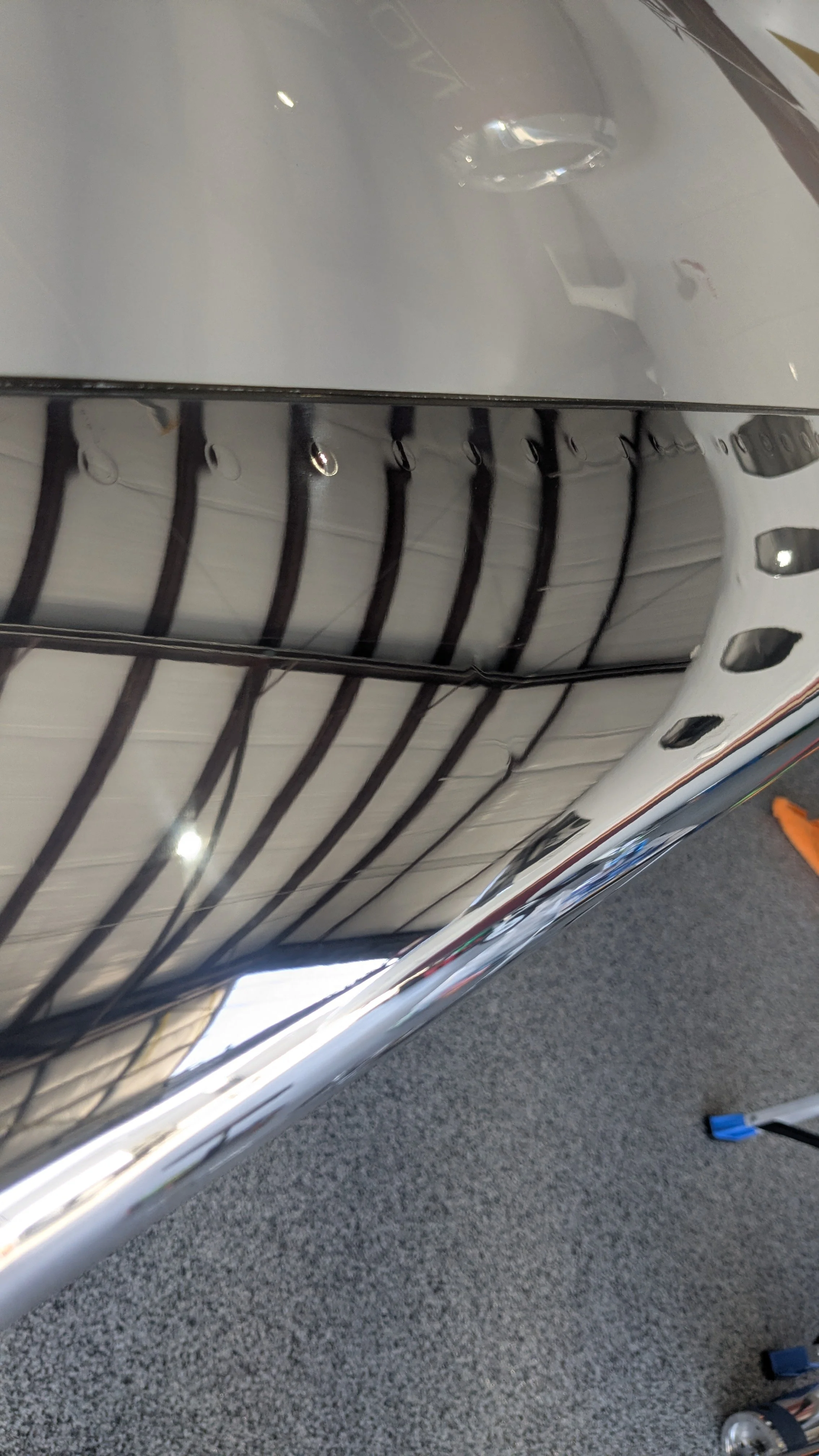 A close-up view of a shiny, reflective surface with black ribs or grill pattern underneath, possibly part of a vehicle or machinery, on a speckled gray floor.