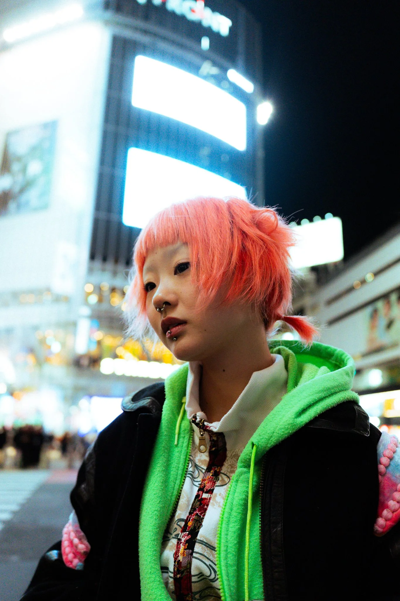 A young woman with bright orange hair featuring bangs and pigtails, wearing a neon green hoodie, black jacket, and white patterned shirt, standing in an urban setting at night with bright city lights and billboards in the background.