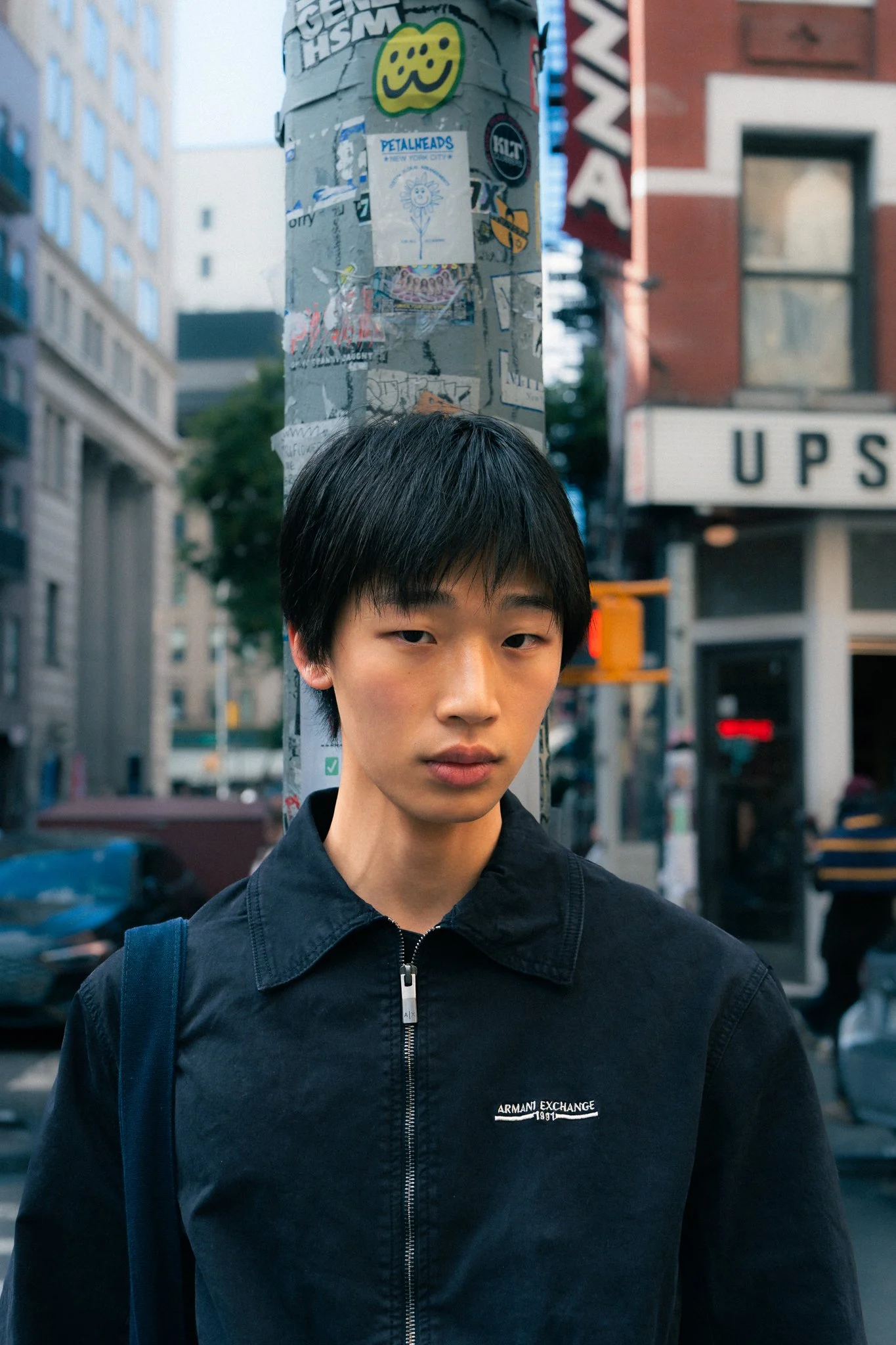 A young man with black hair and fair skin stands outdoors in an urban setting, wearing a black Armani Exchange jacket with a zipper, next to a pole covered in stickers and posters. The background includes buildings, a street, and an 'UPS' sign.