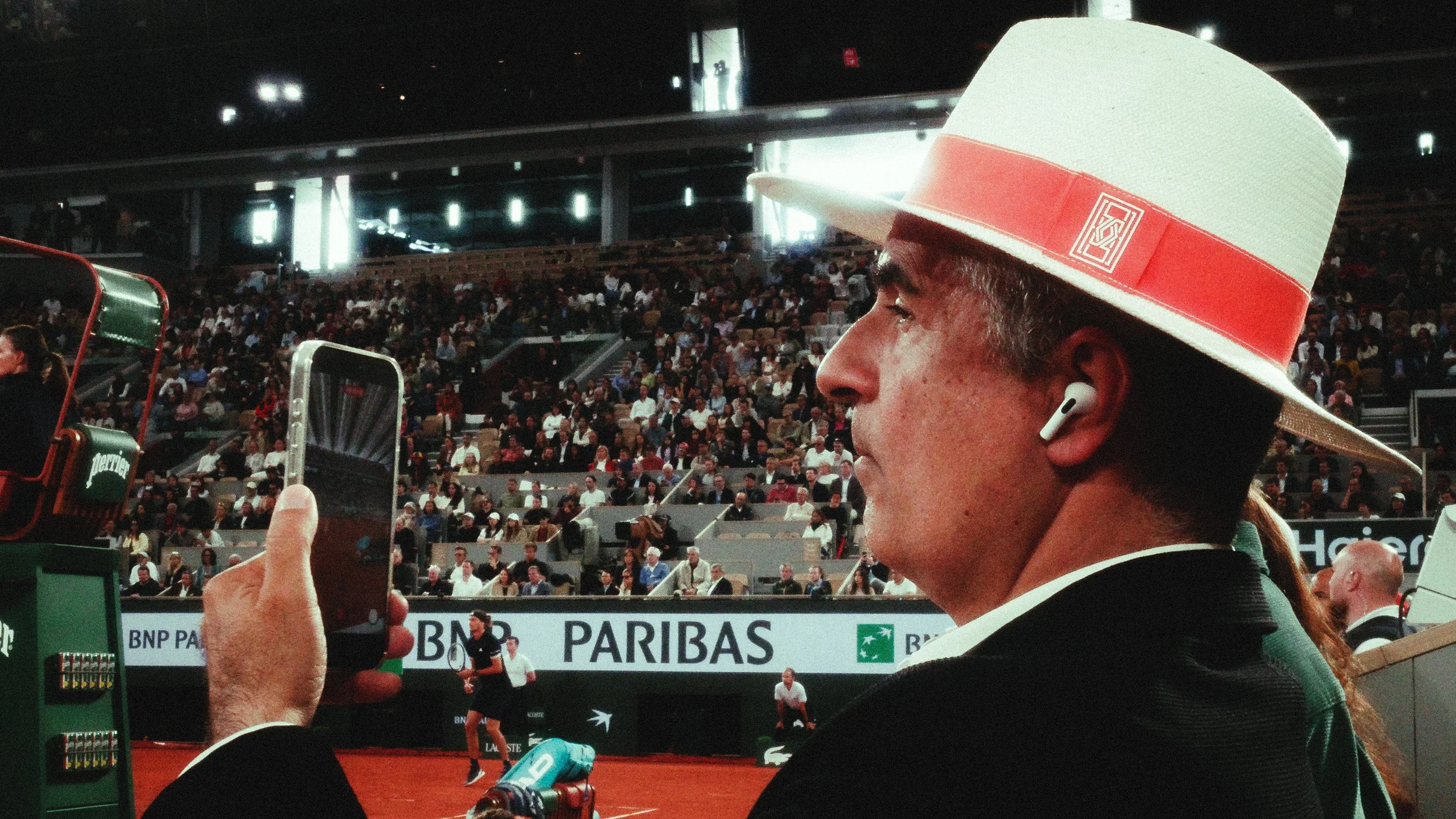 A man wearing a white hat with a red band and AirPods, taking a photo or video at a tennis match in a large indoor stadium filled with spectators.