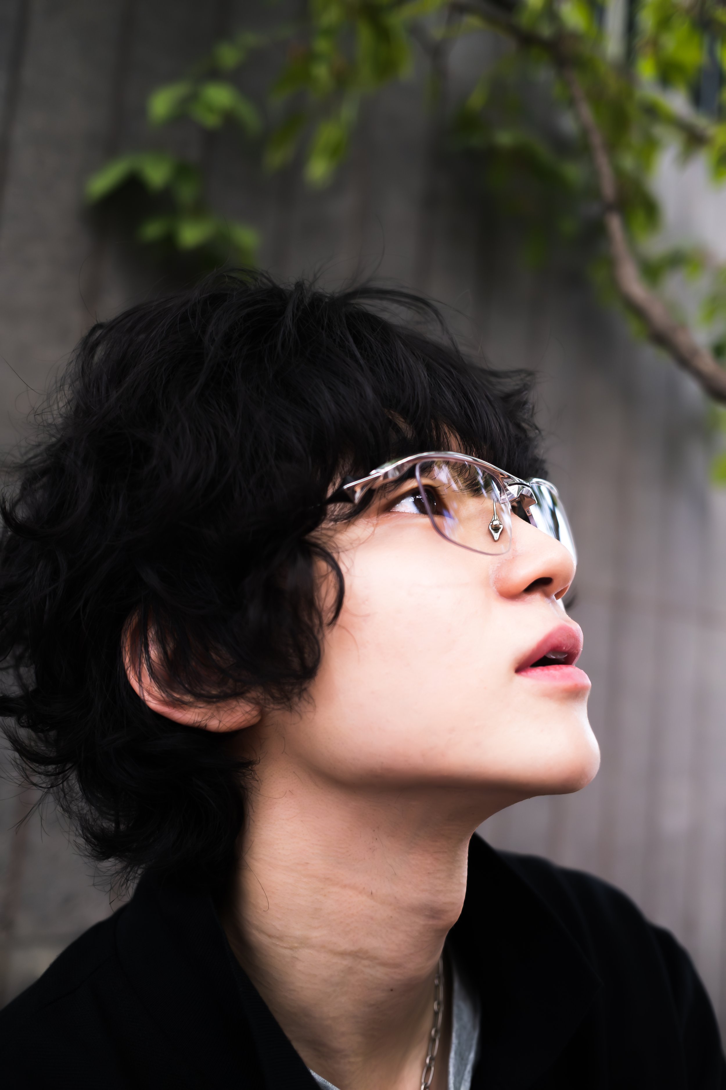 Close-up of a person with short curly black hair wearing glasses, a silver chain necklace, and a black jacket, looking upward with a blurred outdoor background including green leaves and a gray wall.