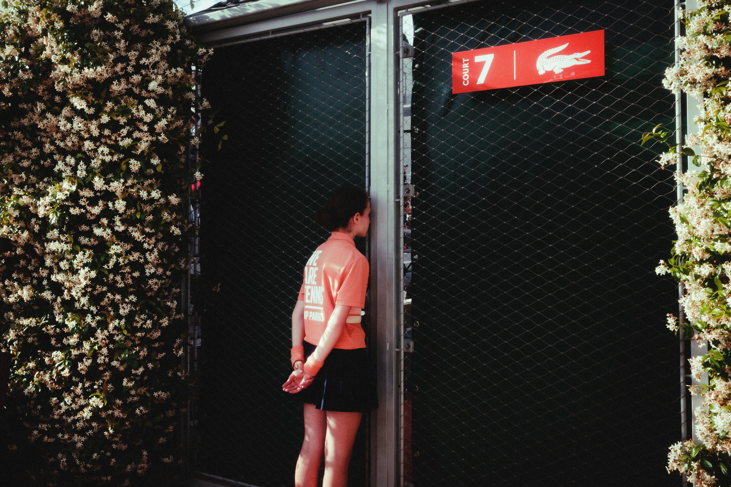 A person with dark hair tied back is standing outside a black gate with a sign reading 'Court 7' and a crocodile logo. The person is wearing a light orange shirt and dark shorts, and is observing the gate. White flowering plants with pink accents are