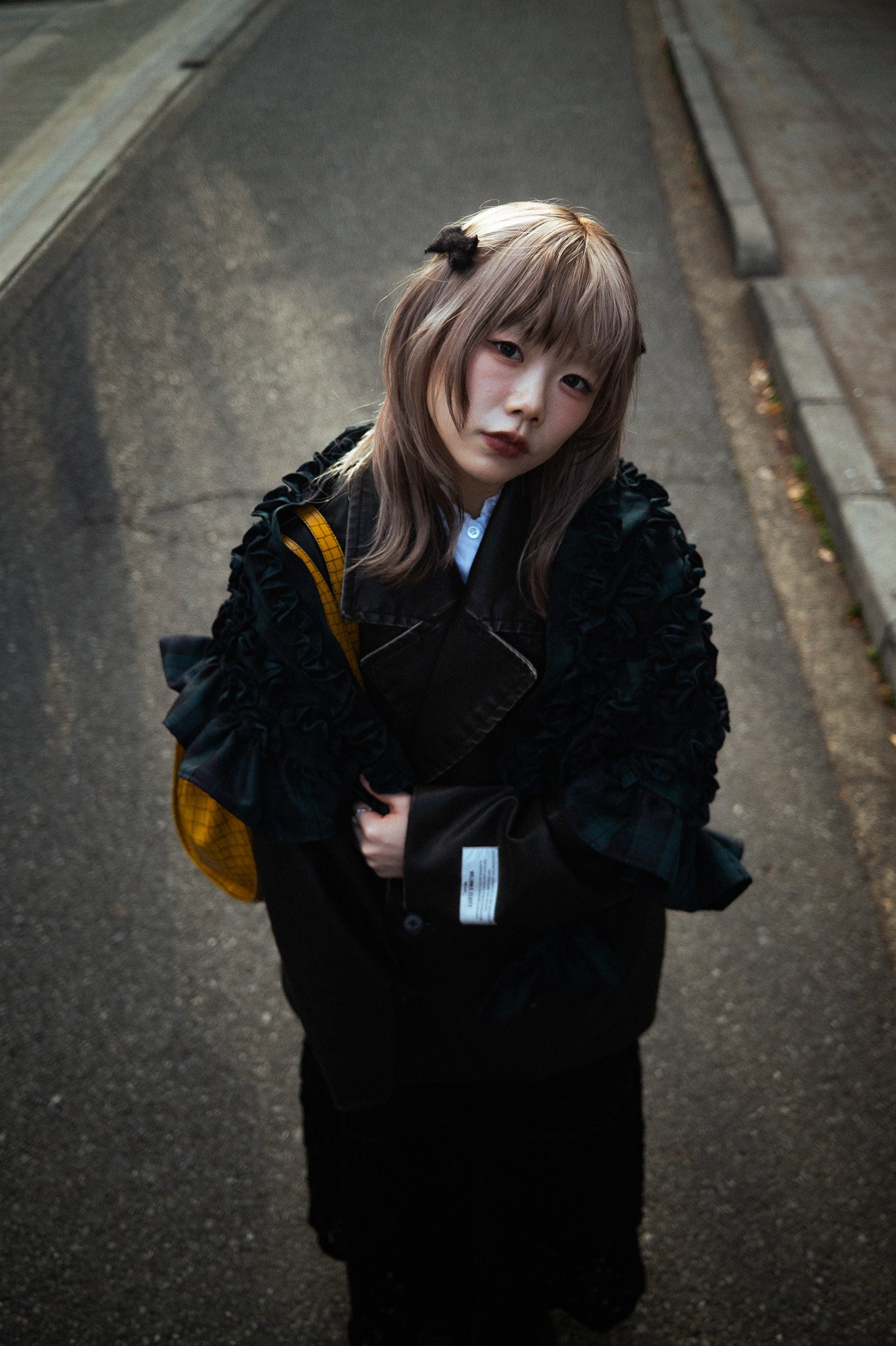 A young woman with shoulder-length light brown hair and bangs, wearing a ruffled black jacket and carrying a yellow checkered backpack, standing on a sidewalk at night with streetlights illuminating the scene.