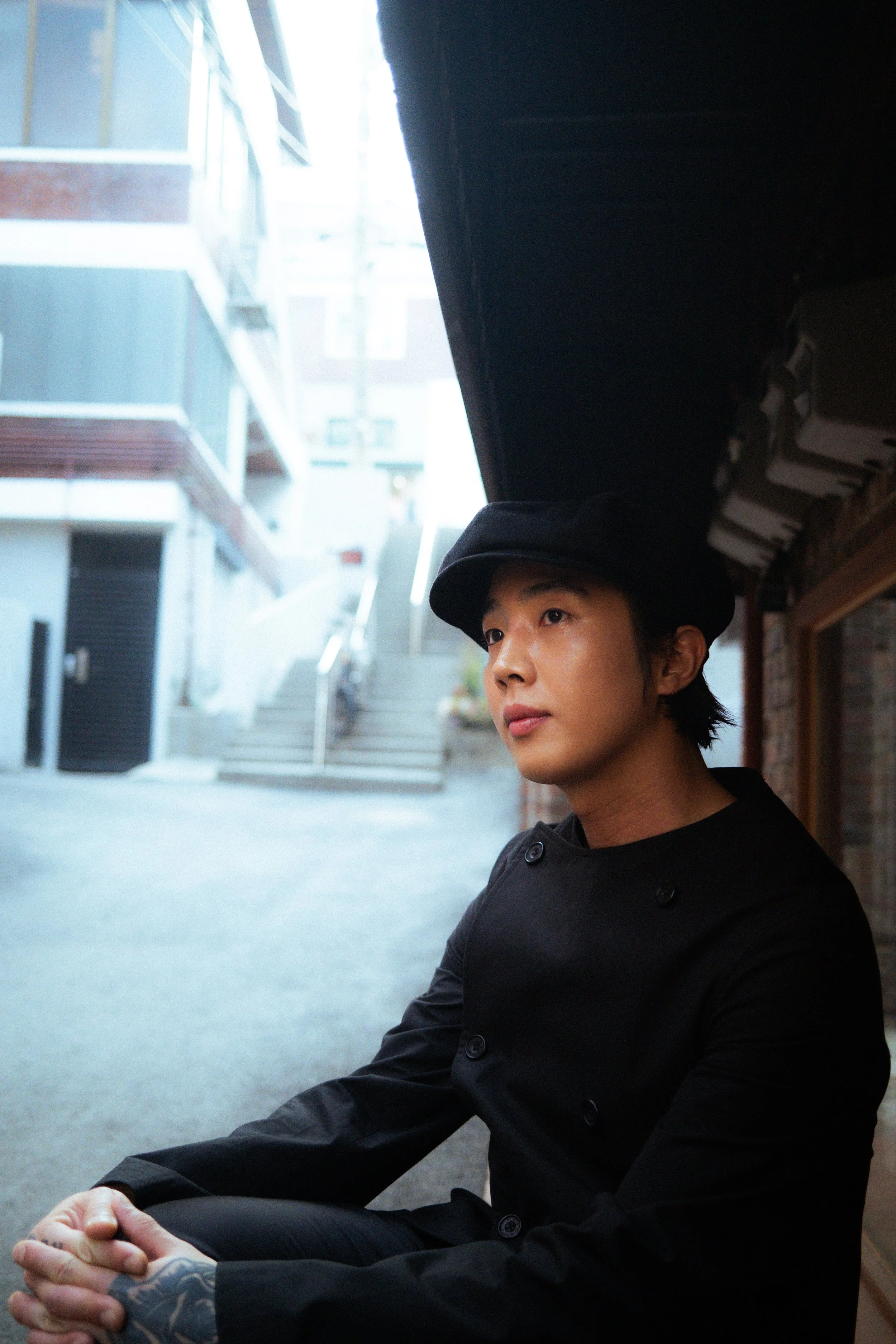 A person with a tattooed hand wearing a black coat and a black hat, sitting outdoors near a building with stairs and windows, looking to the side.