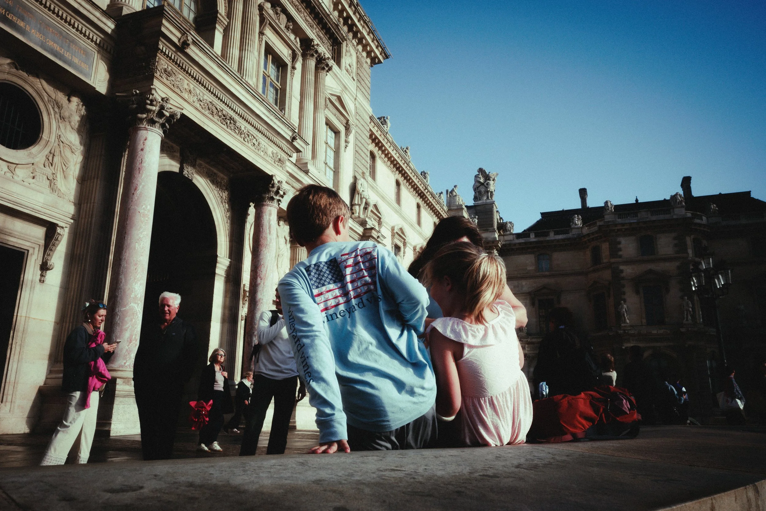 People gathered outside a historic building with ornate architecture, including pillars and sculptures, during daytime with clear blue sky.