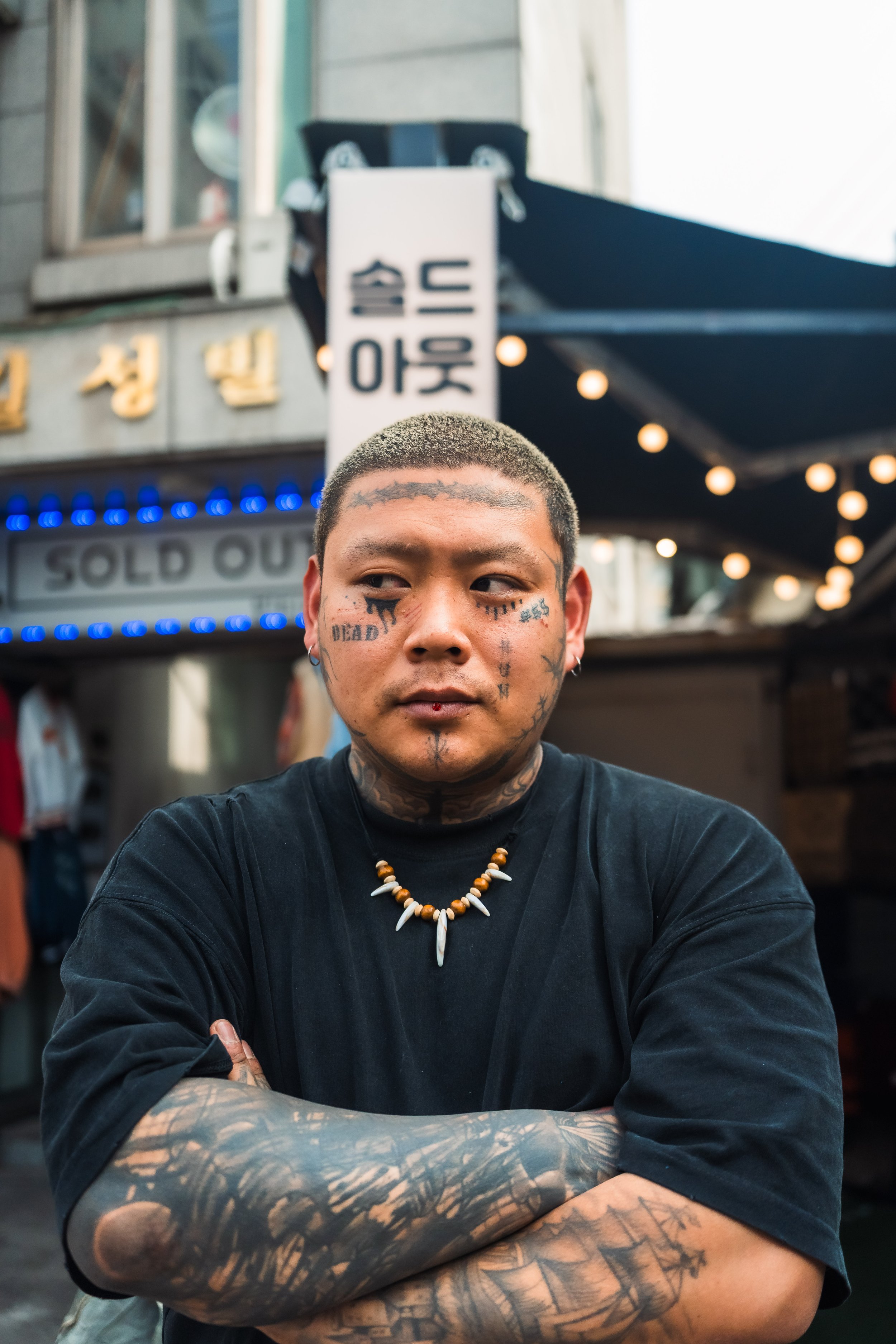 A young man with tattoos on his face and arms, wearing a black shirt and a beaded necklace, standing outdoors with his arms crossed, in front of a building with a sign containing Korean characters and blue lights.