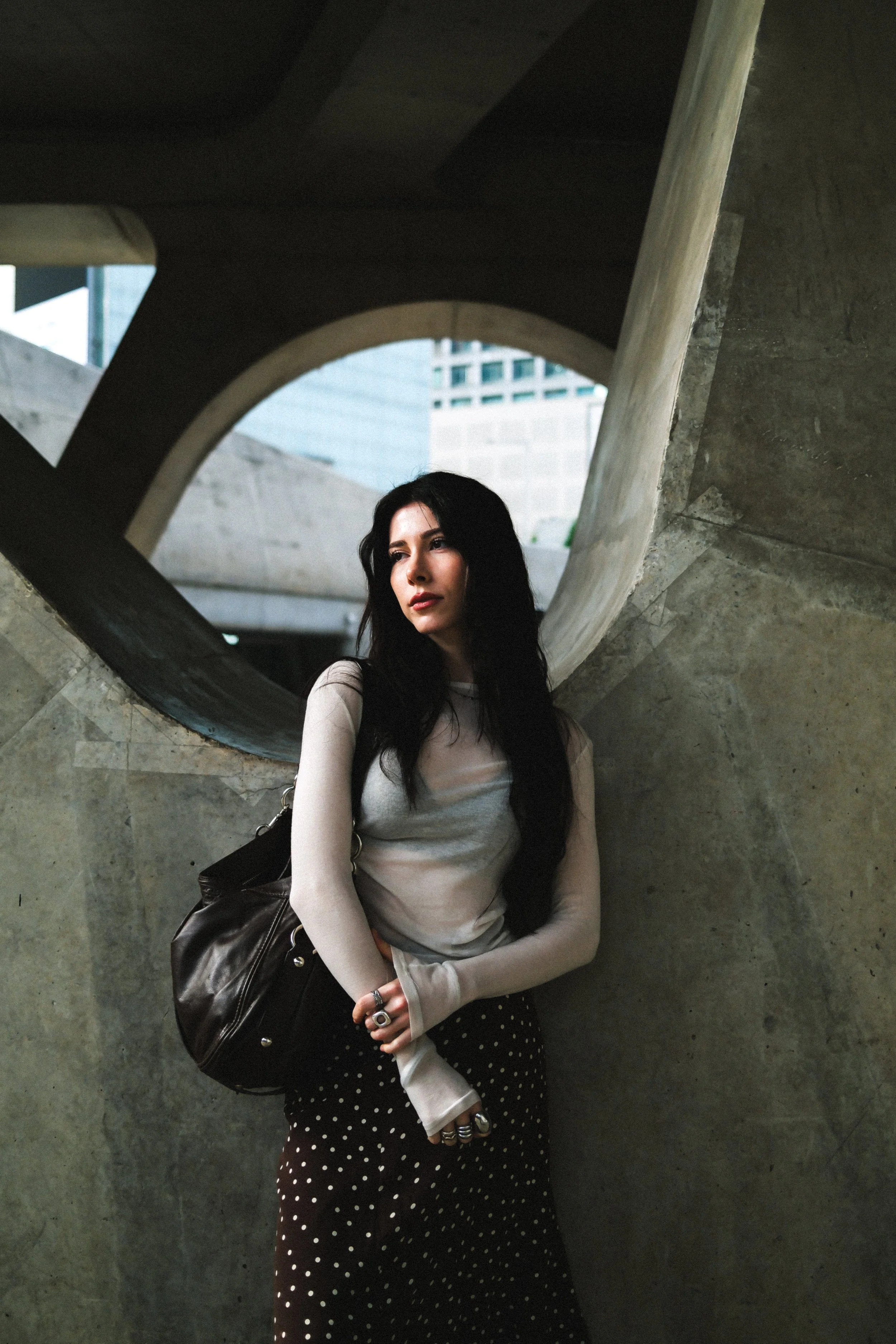 A woman with long dark hair standing under a concrete structure with circular and angular shapes, wearing a light-colored long-sleeve top, polka-dot skirt, and carrying a black bag.