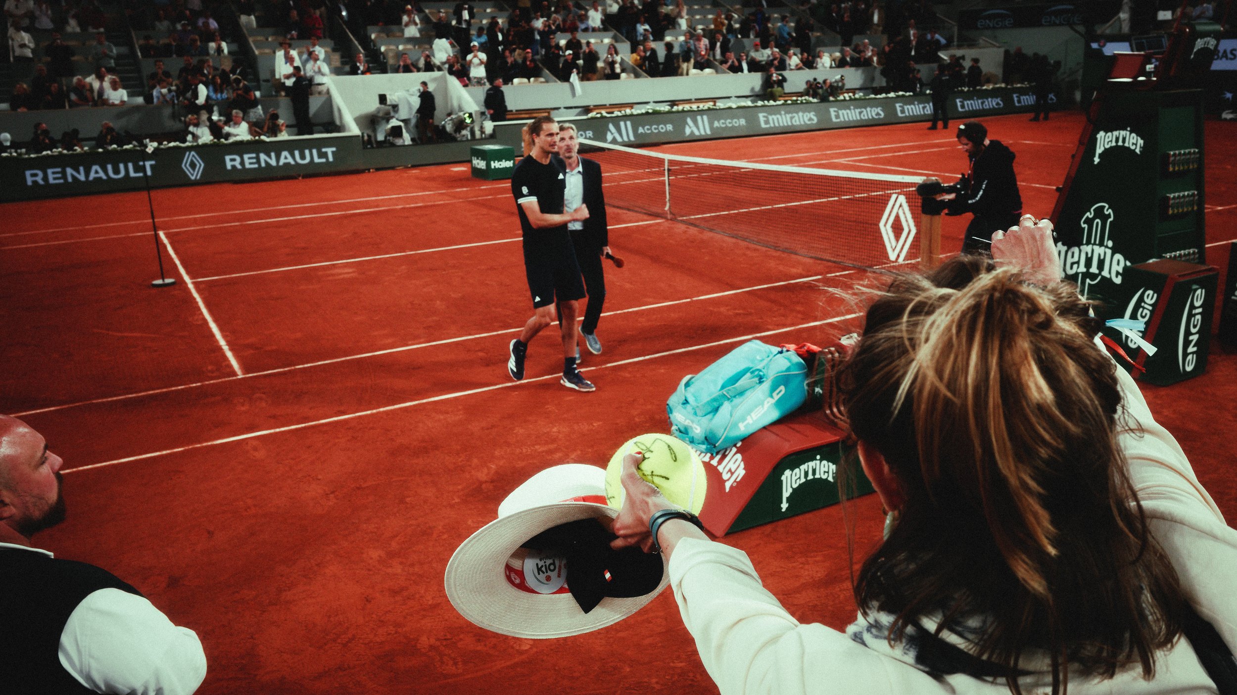 A tennis player walking on a clay court after a match, with a woman in the foreground holding a yellow tennis ball and a hat, and spectators seated around the court.