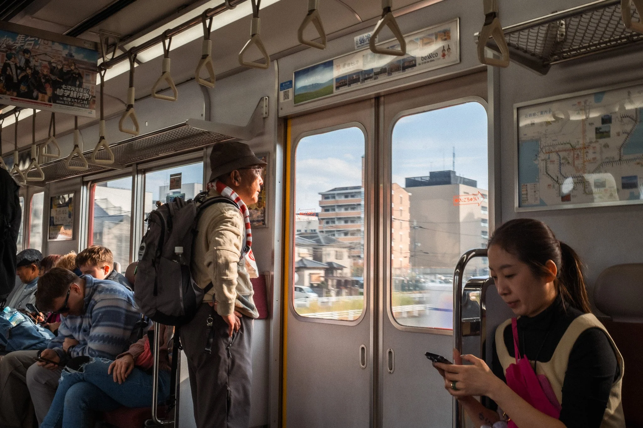 Inside a train, several passengers seated and standing, with city buildings visible through the windows.