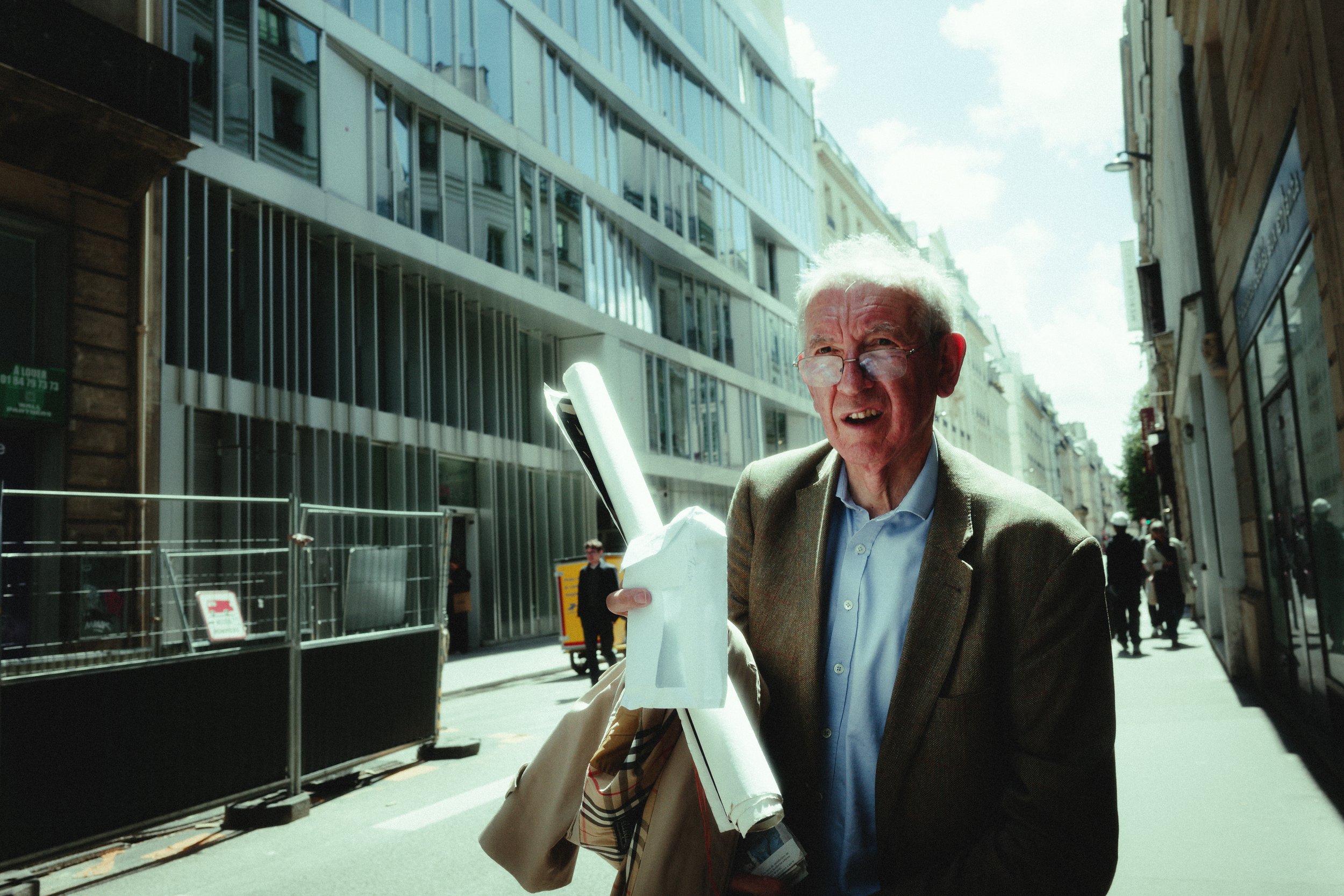 An elderly man with white hair and glasses walking on a city street holding a rolled-up poster and a bag. Behind him are tall modern buildings and other pedestrians.