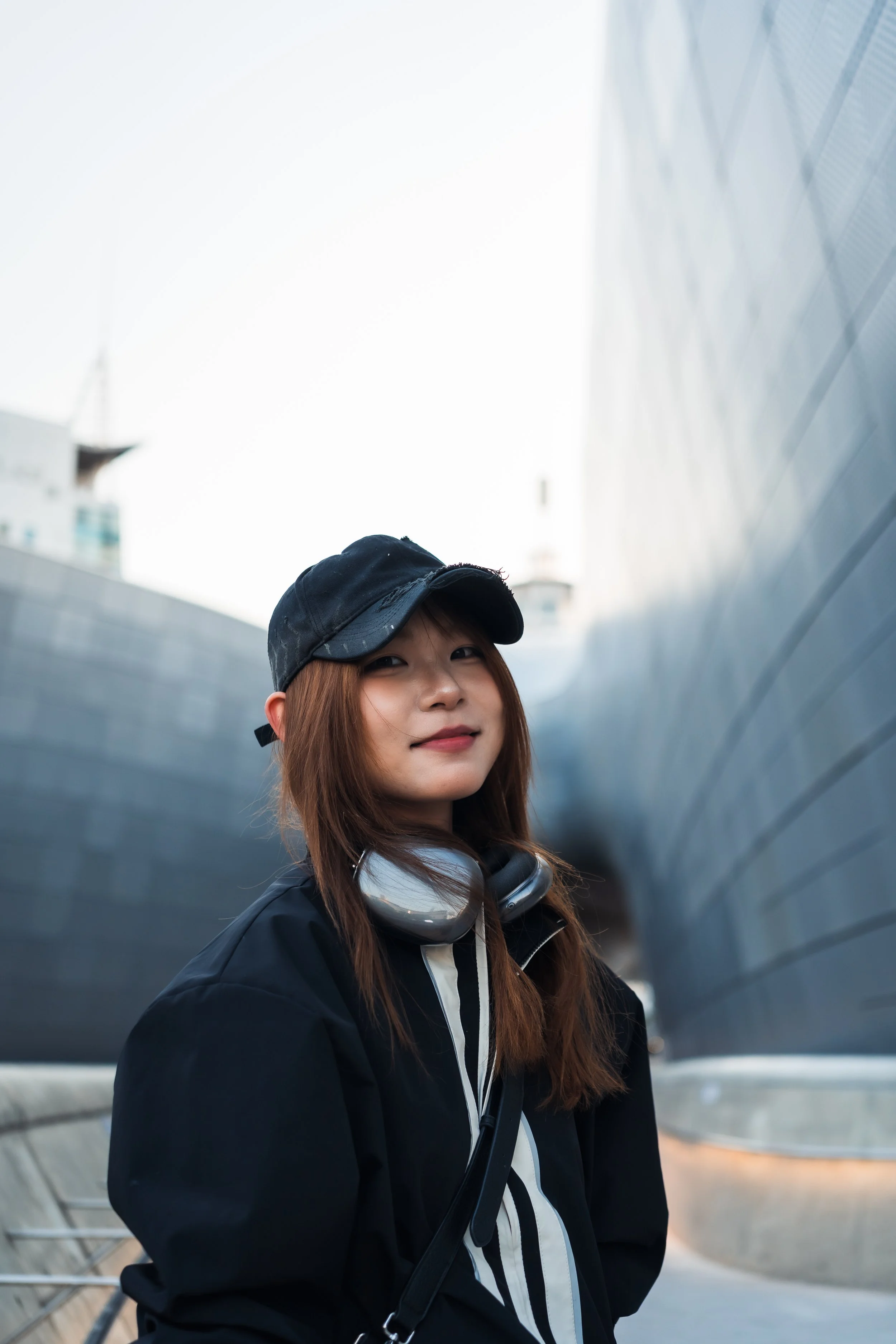 Young woman with brown hair wearing a black cap, black jacket, and headphones around her neck, standing outdoors with modern architectural structures in the background.