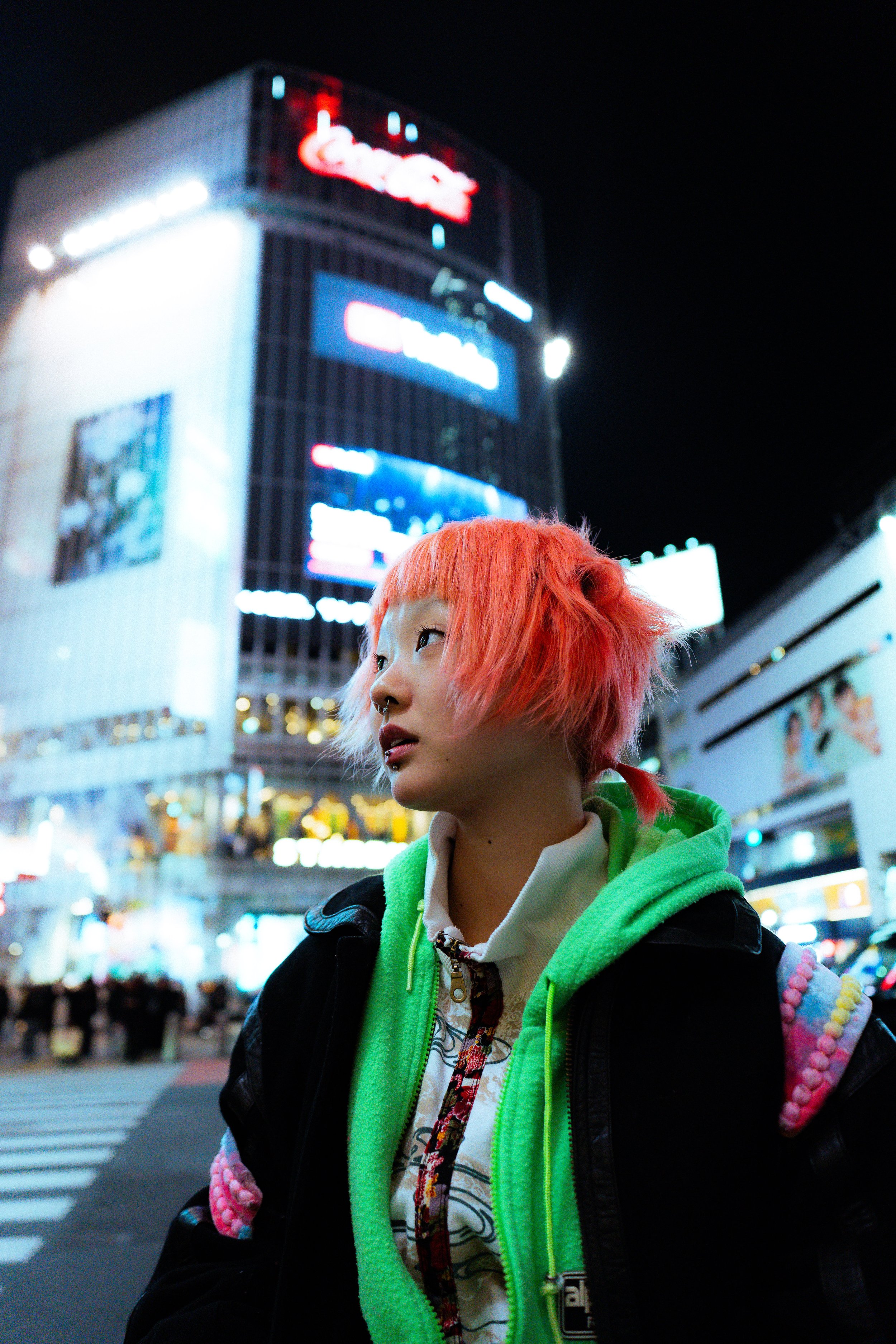 A young person with bright orange hair, piercing, and lip rings, standing on a city street at night with illuminated billboards and crowded streets in the background.