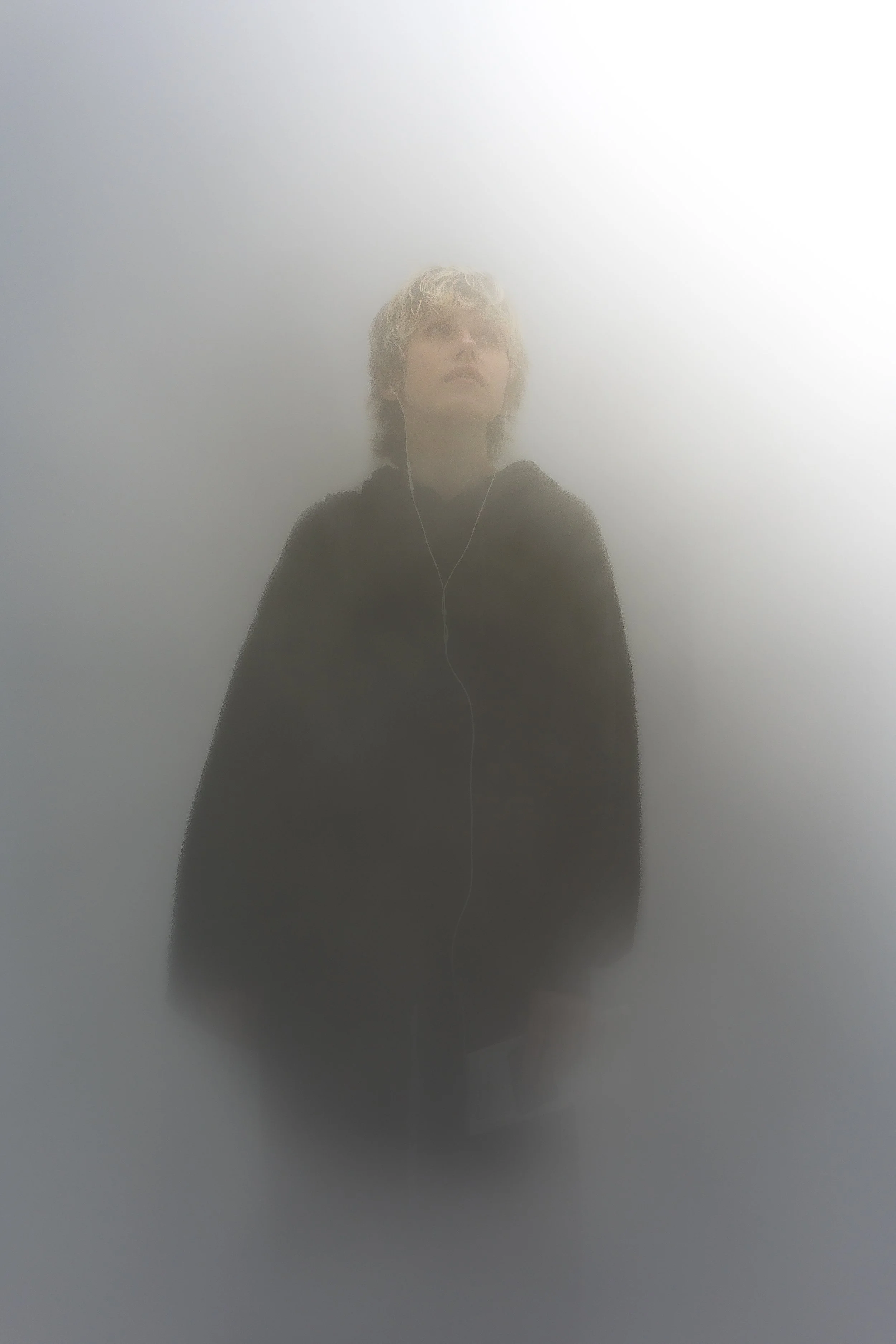 Person with short, wavy blonde hair wearing a black hoodie, listening to headphones, standing against a foggy or misty background.