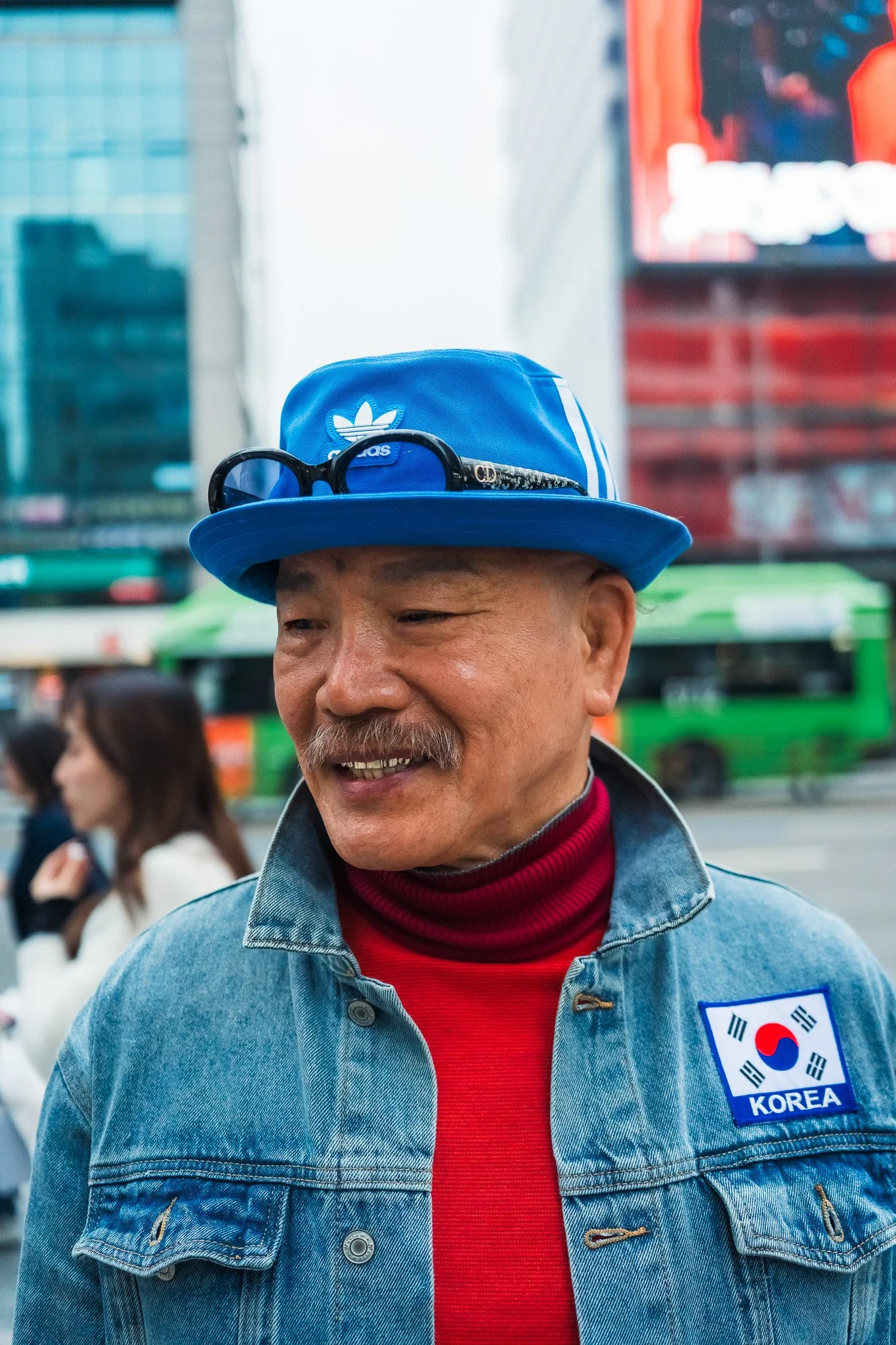 A man in a blue denim jacket with a South Korea patch, red turtleneck, and blue Adidas hat with sunglasses, smiling outdoors in a city with people and tall buildings in the background.