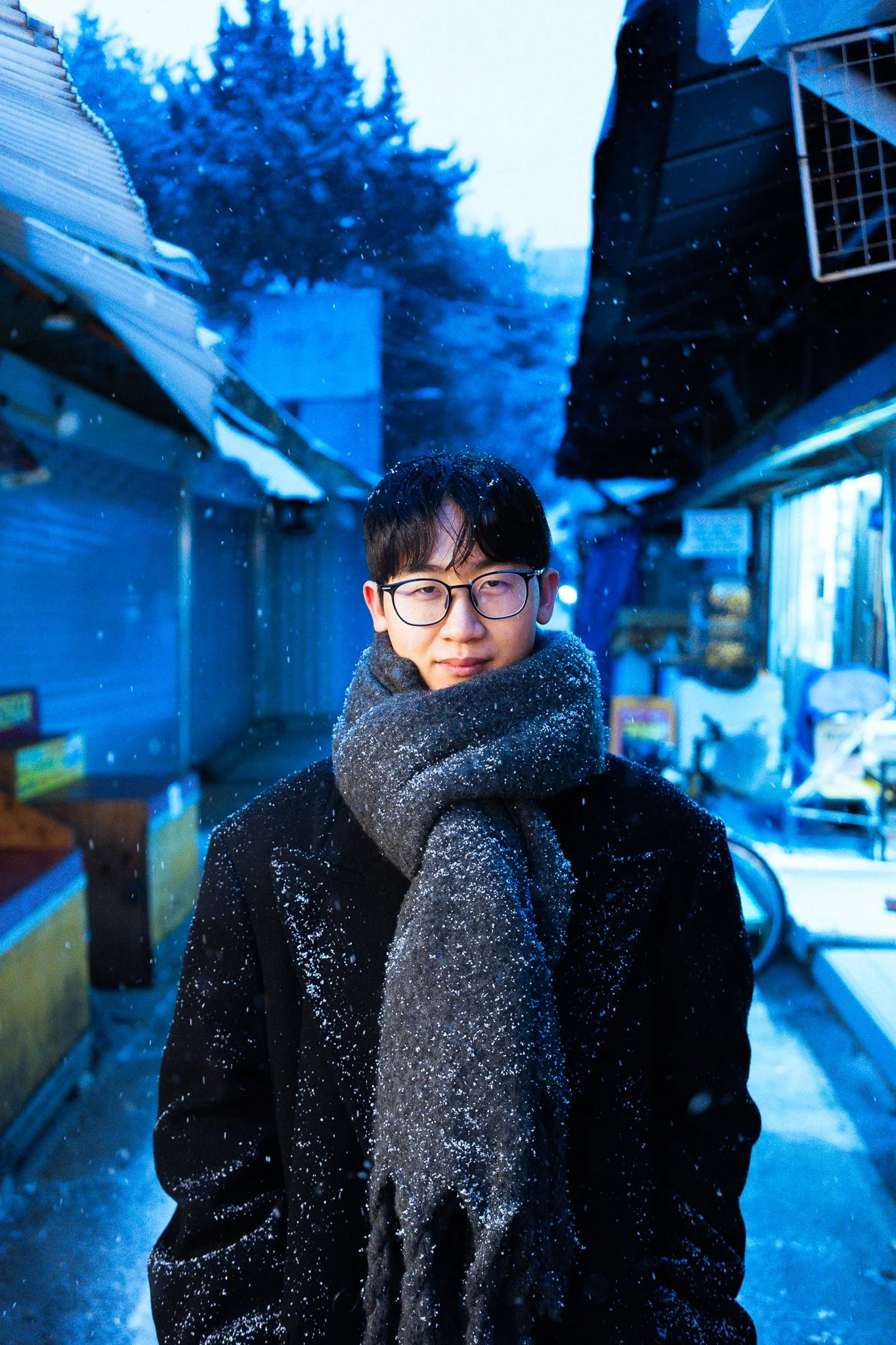 Young man with glasses and dark hair standing outdoors in a snowy alleyway, wearing a scarf and black coat, with snow falling around him and trees in the background.