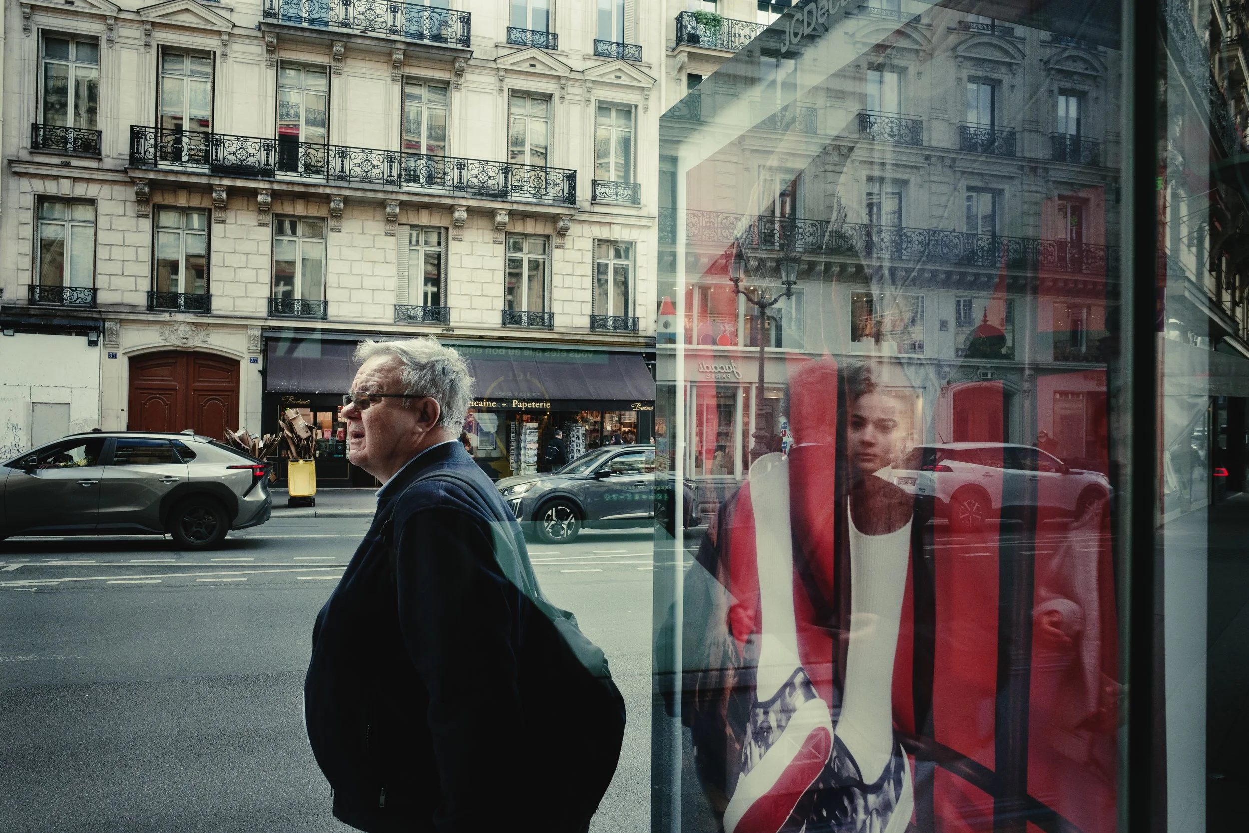 Street scene in Paris with a historic building featuring ornate balconies. A man with gray hair, glasses, and a dark jacket walks past, with reflections of a young man and store signage in the glass window.