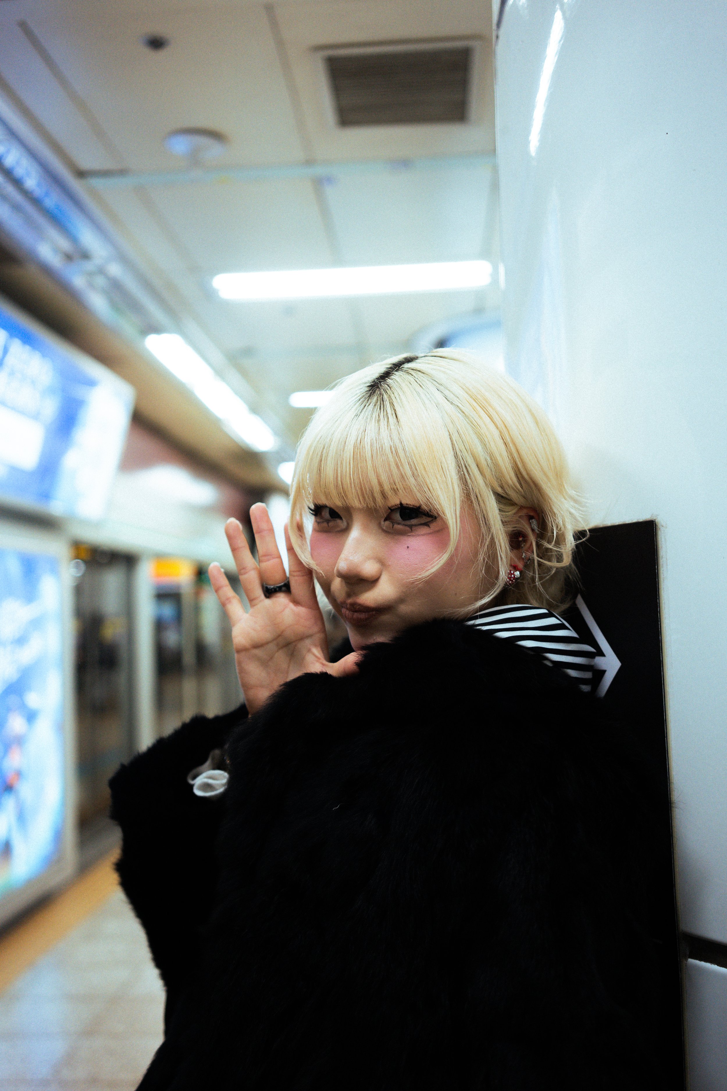 A person with blonde hair and eye makeup is posing in front of a wall in a subway or train station, smiling and waving with their hand. They are wearing a black furry coat.