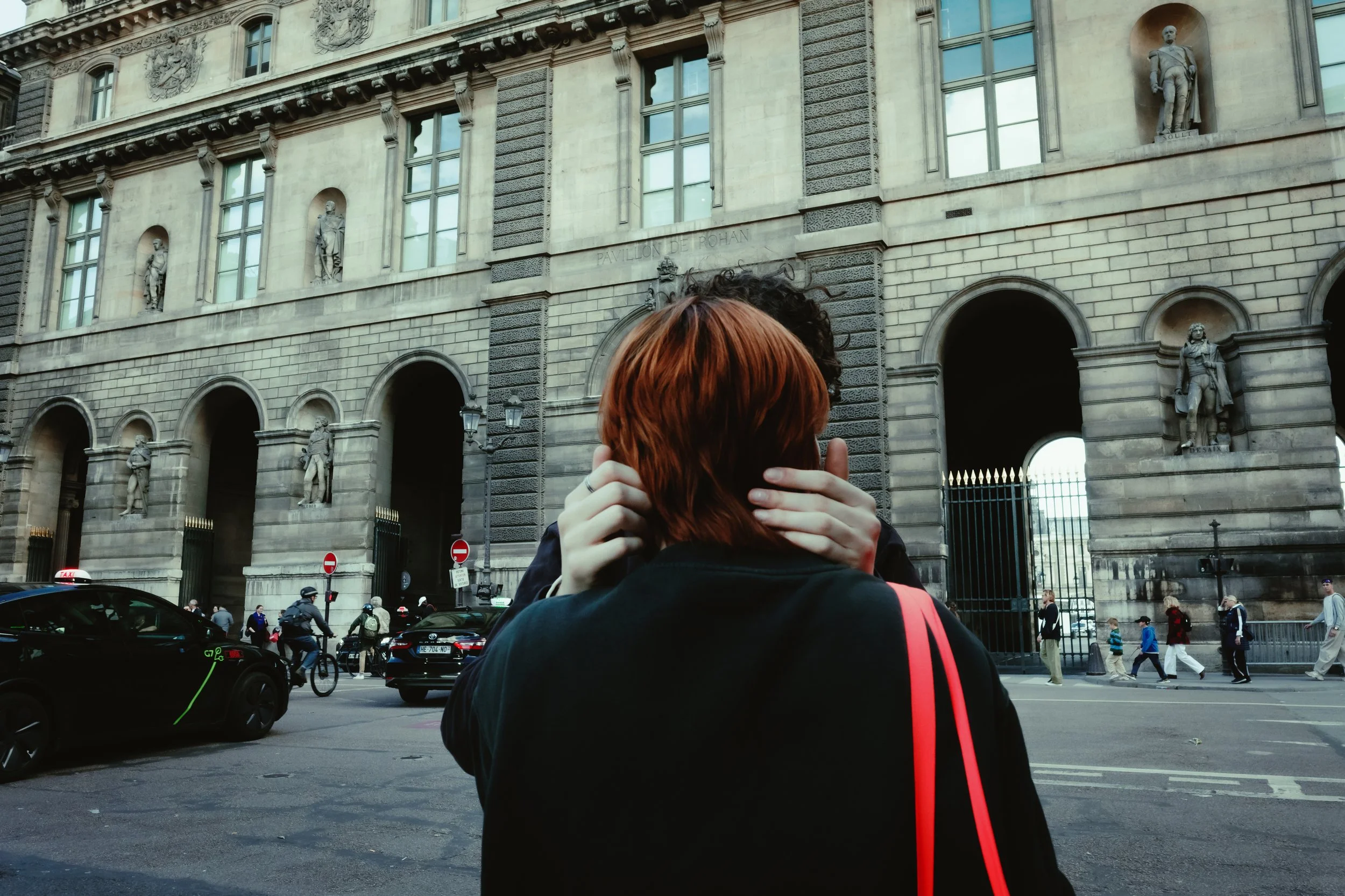 A person with reddish hair embracing someone outside in front of an ornate historic building with statues and arches, with cars and pedestrians on the street.