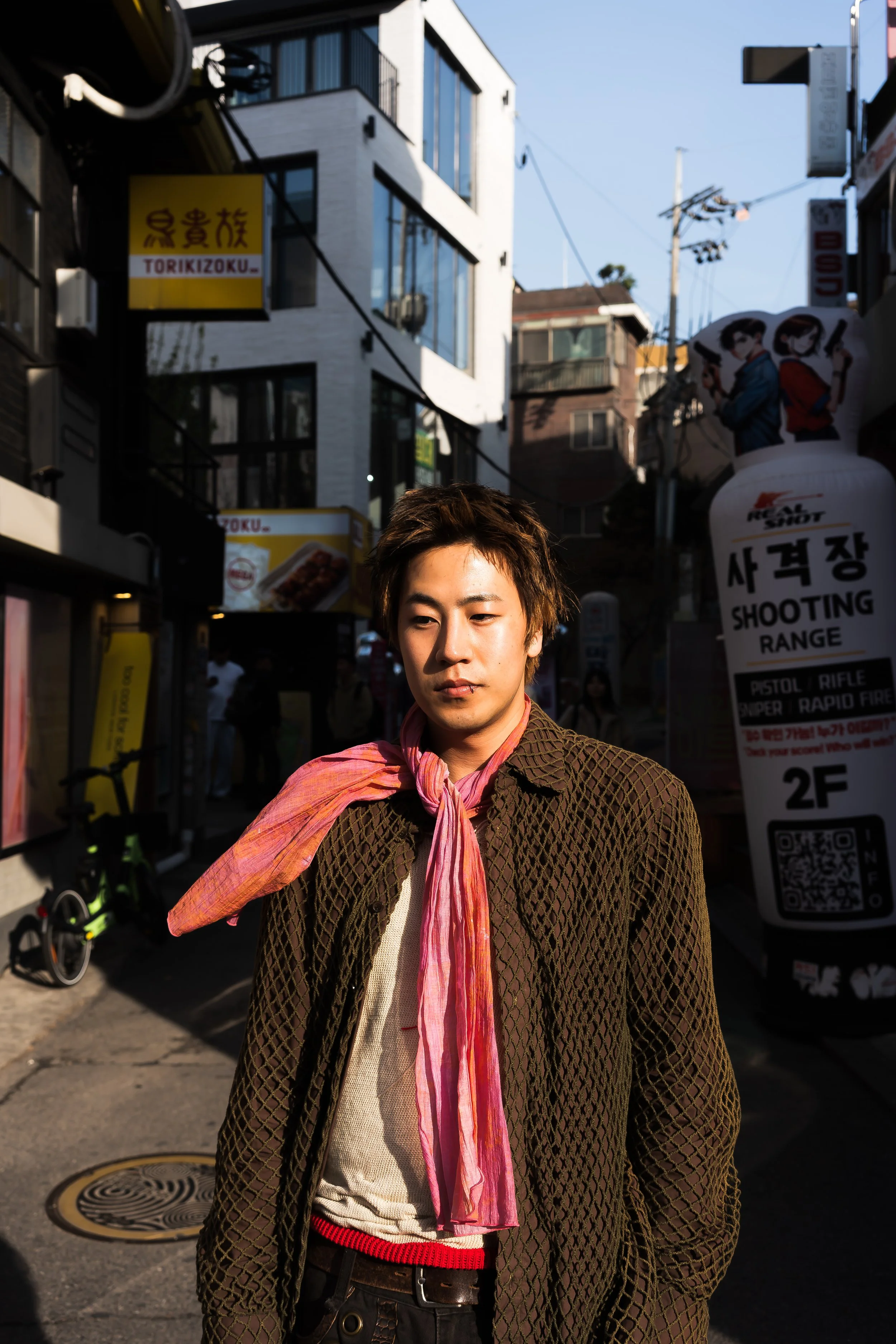 Young man with fair skin and brown hair, wearing a patterned brown coat, beige shirt, black pants with a red belt, and a pink scarf tied around his neck, standing on a city street with signs, buildings, and utility poles in the background.